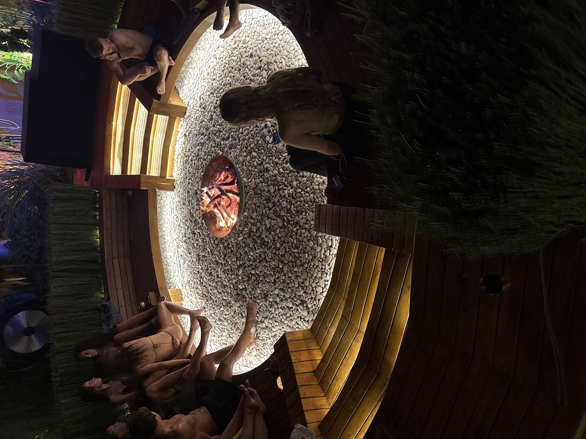 The image depicts an evening scene at an outdoor leisure area in Kuta Selatan, Indonesia, featuring a central fire pit surrounded by tiered wooden seating. Approximately nine individuals are present, predominantly young adults, engaged in relaxation.
In the center, a circular metal fire pit emits bright orange flames and glowing embers, resting on an illuminated bed of small, smooth, light-grey pebbles. Encircling this area is a dark wooden structure comprising benches and steps, lit from underneath by warm, yellow strip lights.
On the left side of the seating area, several individuals are visible. One male, shirtless and wearing dark shorts, sits with bare feet on the lowest tier. Beside him, multiple females are seated, some in swimwear or light attire, with bare feet, appearing to be conversing. One woman wears a light-brown bikini top and bottoms.
On the upper right section of the wooden seating, a male individual, shirtless and wearing dark shorts, sits cross-legged with bare feet. He holds a hand to his ear, possibly using a phone or adjusting an earbud. To his right, partially obscured, are bare feet of another person.
In the foreground, at the bottom right, a female with long, light-brown hair, viewed from behind, sits on a lower section of the wooden structure or a cushion, wearing a light-colored halter top and dark trousers. She is actively viewing a smartphone.
The perimeter of the area features tall, slender green grasses or reeds, subtly illuminated. In the far background, dark structures are visible, including a large, metallic, disc-shaped object mounted vertically, and a large, dark rectangular object, possibly an equipment cover. The overall lighting suggests nighttime, with artificial sources illuminating the central gathering space.