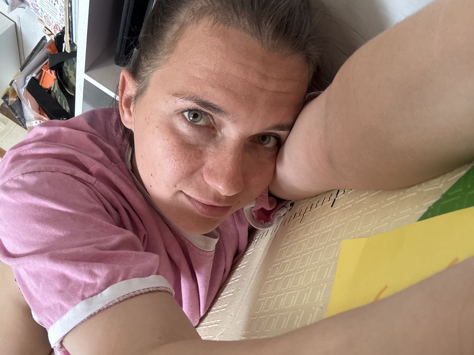 A woman with fair skin, green eyes, and brown hair pulled back is lying down in what appears to be a room with white shelves and various items on them. She is wearing a pink t-shirt with white trim. Her left arm is bent and positioned under her head. A piece of yellow paper is visible on the bed near her arm. The location in Раздолье, Russia, is not discernible from the image.