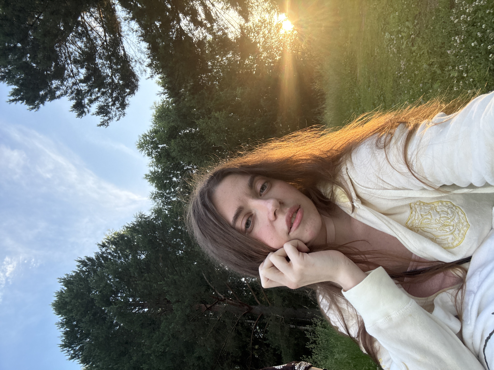 A young woman with long, brown hair is shown in a selfie. She is wearing a light-colored hooded jacket over a pink top. Her left hand is raised to her face, and she is looking directly at the camera. The background consists of trees and a grassy field, with the sun setting behind the trees, casting a warm glow and lens flares across the image. The location is described as Unknown, Unknown.