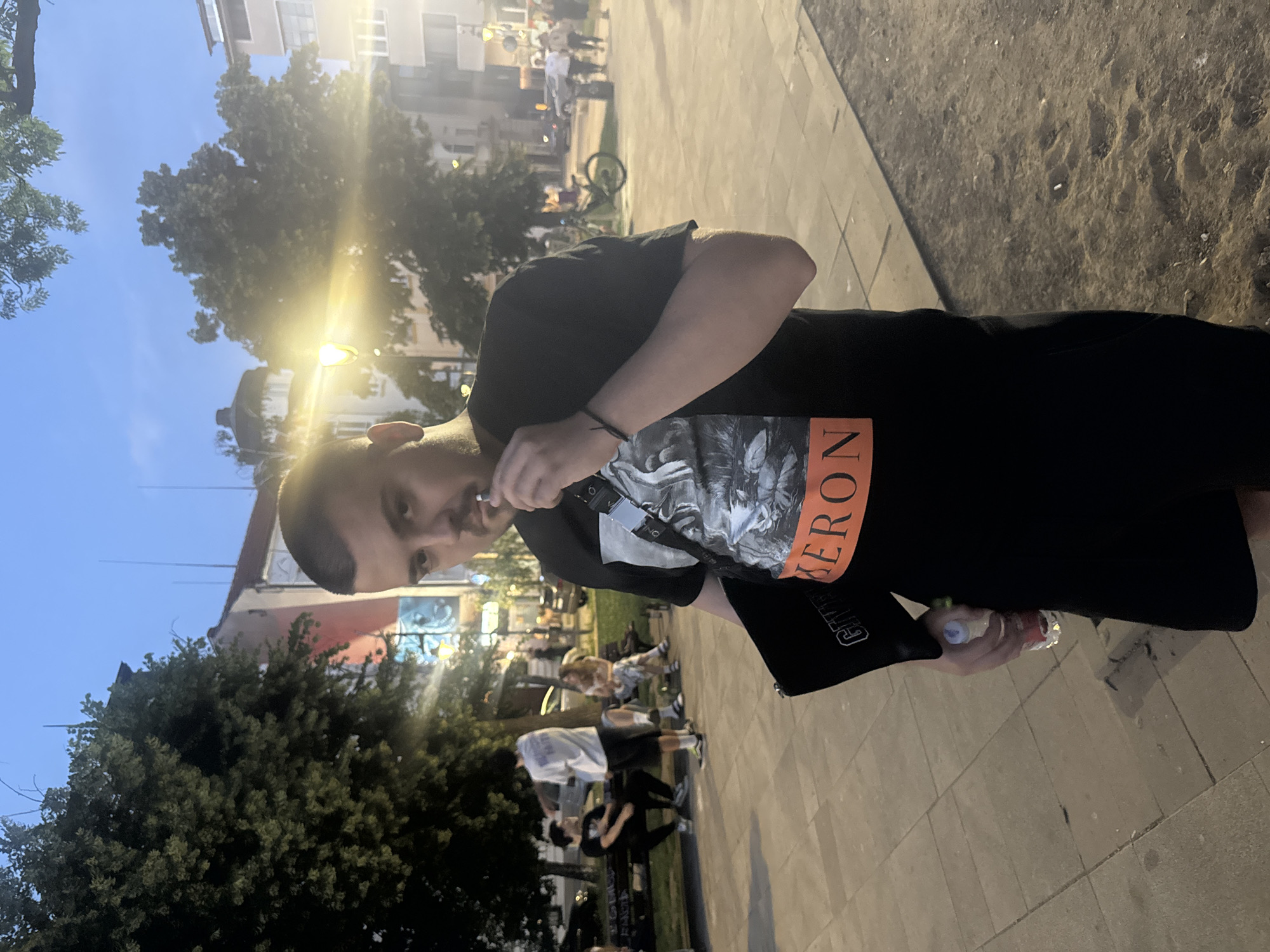 The image depicts a young man standing on a paved area in what appears to be a public urban space, possibly a park or plaza, in Sofia, Bulgaria, as suggested by the provided context. The lighting indicates it is dusk or early evening. The man is wearing a black t-shirt with a graphic print and an orange band across the chest, a black bag slung over his shoulder, and dark shorts. He is holding a cigarette to his lips with his right hand and a bottle of water in his left hand. In the background, trees line the paved area, and buildings with some lights are visible. Several other individuals are present in the background, some sitting on benches and others walking. A bicycle is also visible in the middle ground. The ground surface consists of both paved tiles and areas of dirt or sand.