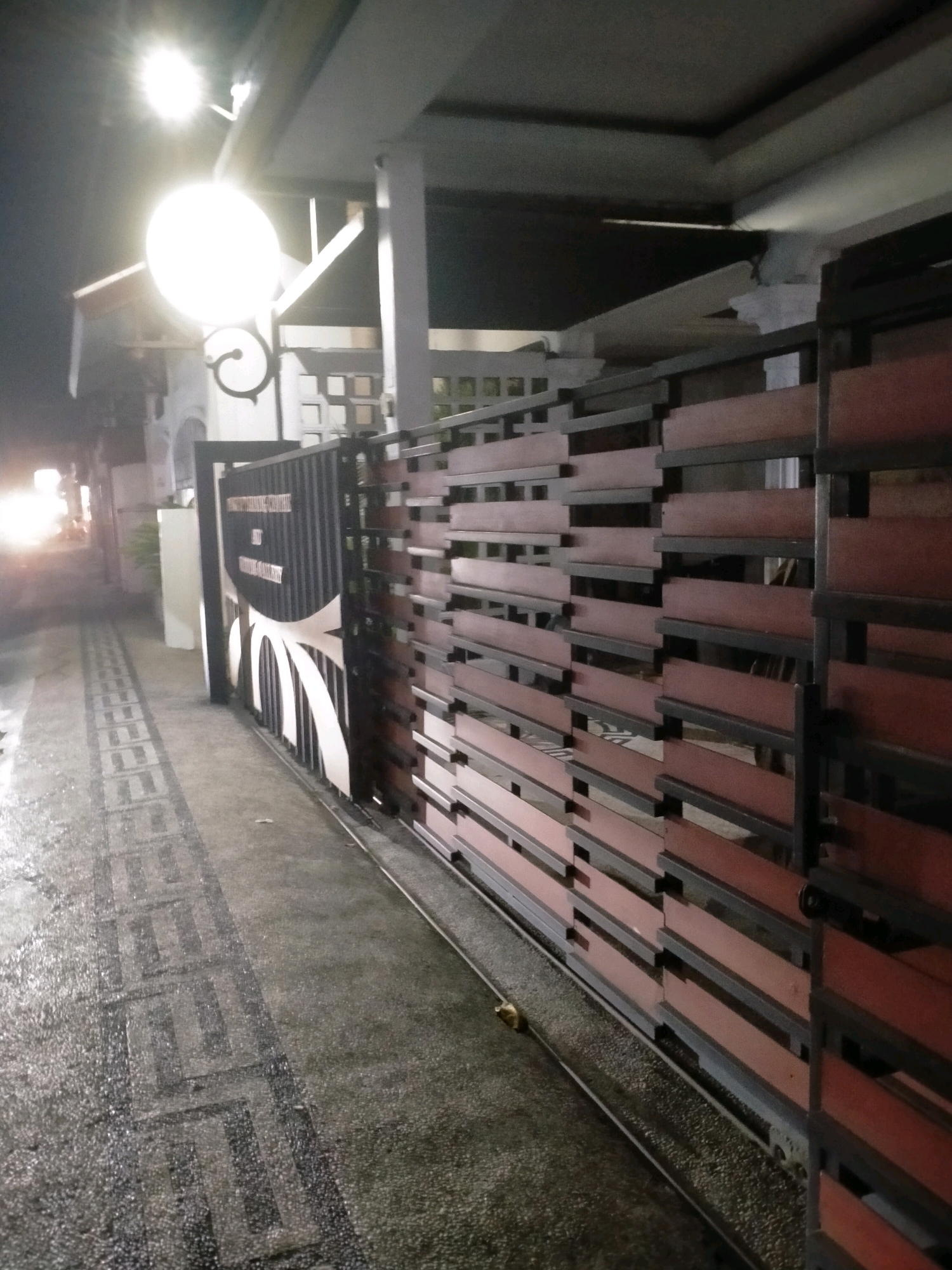 The image shows a night-time view of an open gate and fence, likely a residential property, on a street in Kasihan, Indonesia. The gate, made of black metal with decorative white arches, is partially open, revealing a glimpse of what's inside. The fence to the right is constructed with horizontal wooden slats in a reddish-brown color, interspersed with black metal supports. The ground is a paved walkway with a patterned mosaic design, appearing wet, suggesting recent rain or dampness. A bright outdoor light fixture with a circular shade is visible overhead, illuminating the scene. In the background, a building with white walls and some windows can be seen. The street is dimly lit, with the headlights of a vehicle partially visible on the left, indicating ongoing traffic. The overall impression is of a quiet street at night, with emphasis on the architectural details of the gate and fence.