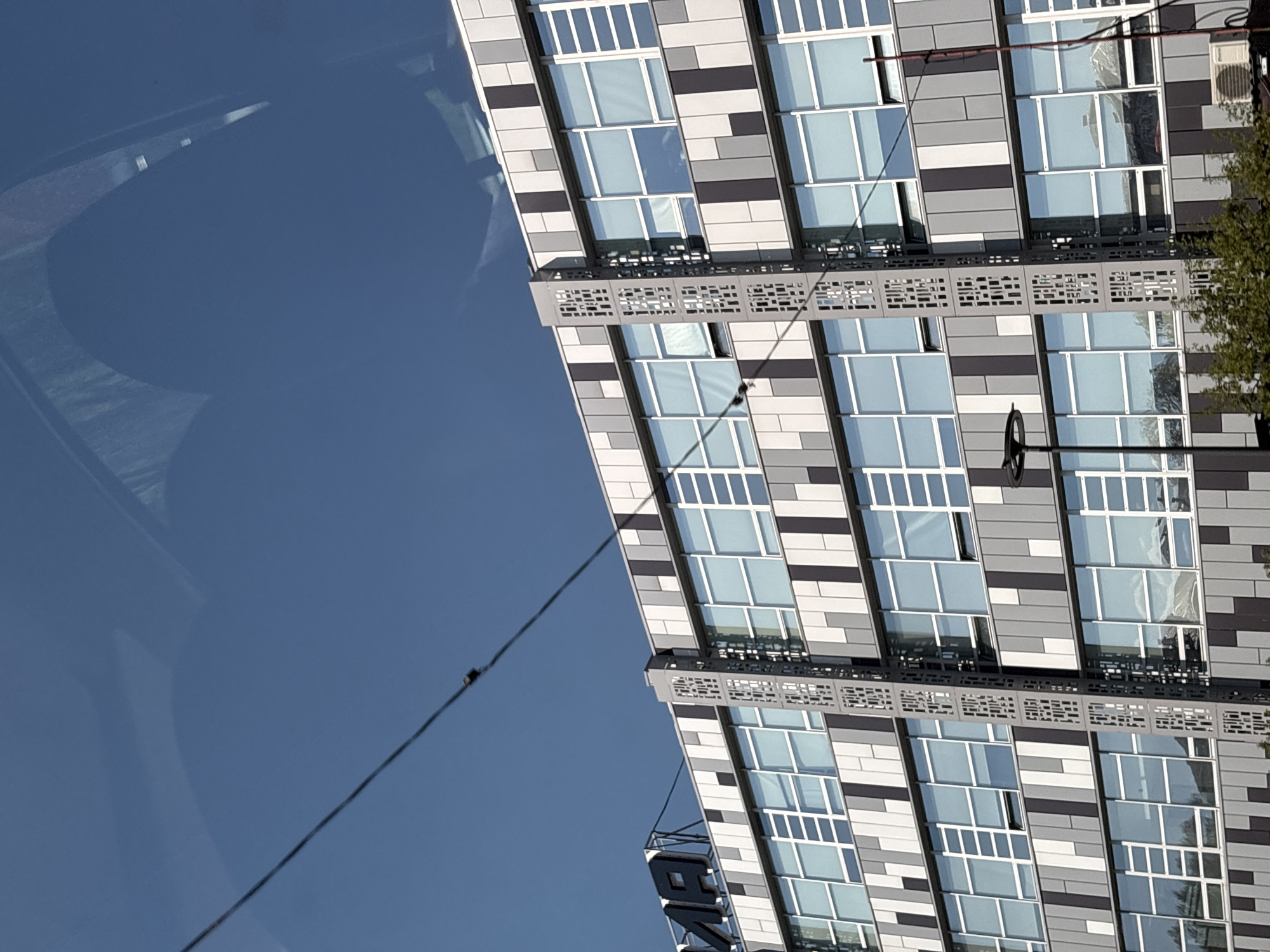 The image shows a portion of a tall building with a modern facade of alternating gray and white rectangular panels and windows. The building's reflective windows mirror the sky. In the lower left of the image, a blue sky reflection is visible, with a thin electrical wire running diagonally across it. The scene is in Perm, Russia, as indicated by the building's design and architectural style.