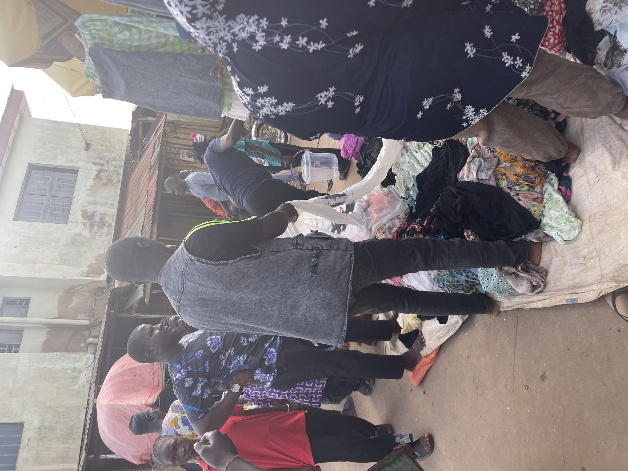 People are gathered at a clothing stall in Akwanga, Nigeria. A pile of clothes sits on a white sheet on the ground. A person in a dark blue shirt with white floral designs and brown pants stands near the clothing pile. A man in a grey vest stands over the clothing, and a person in a blue floral shirt and a person in a red shirt stand beside them. A building is in the background.