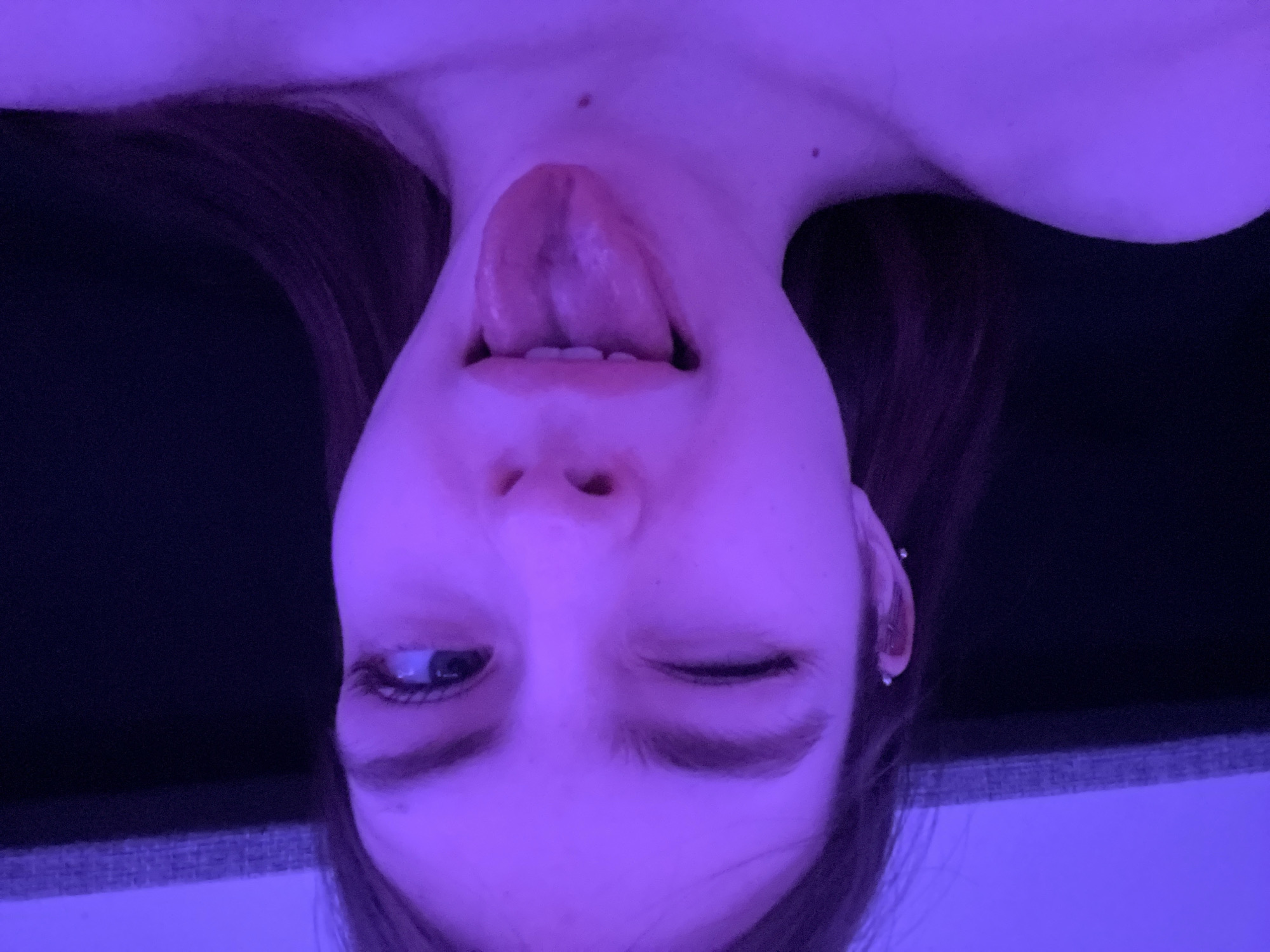 A young woman is positioned upside down, sticking her tongue out. Her right eye is open, and her left eye is closed. The scene is bathed in purple lighting. The background appears to be a dark fabric. There is no indication of the location being Saint Petersburg, Russia.