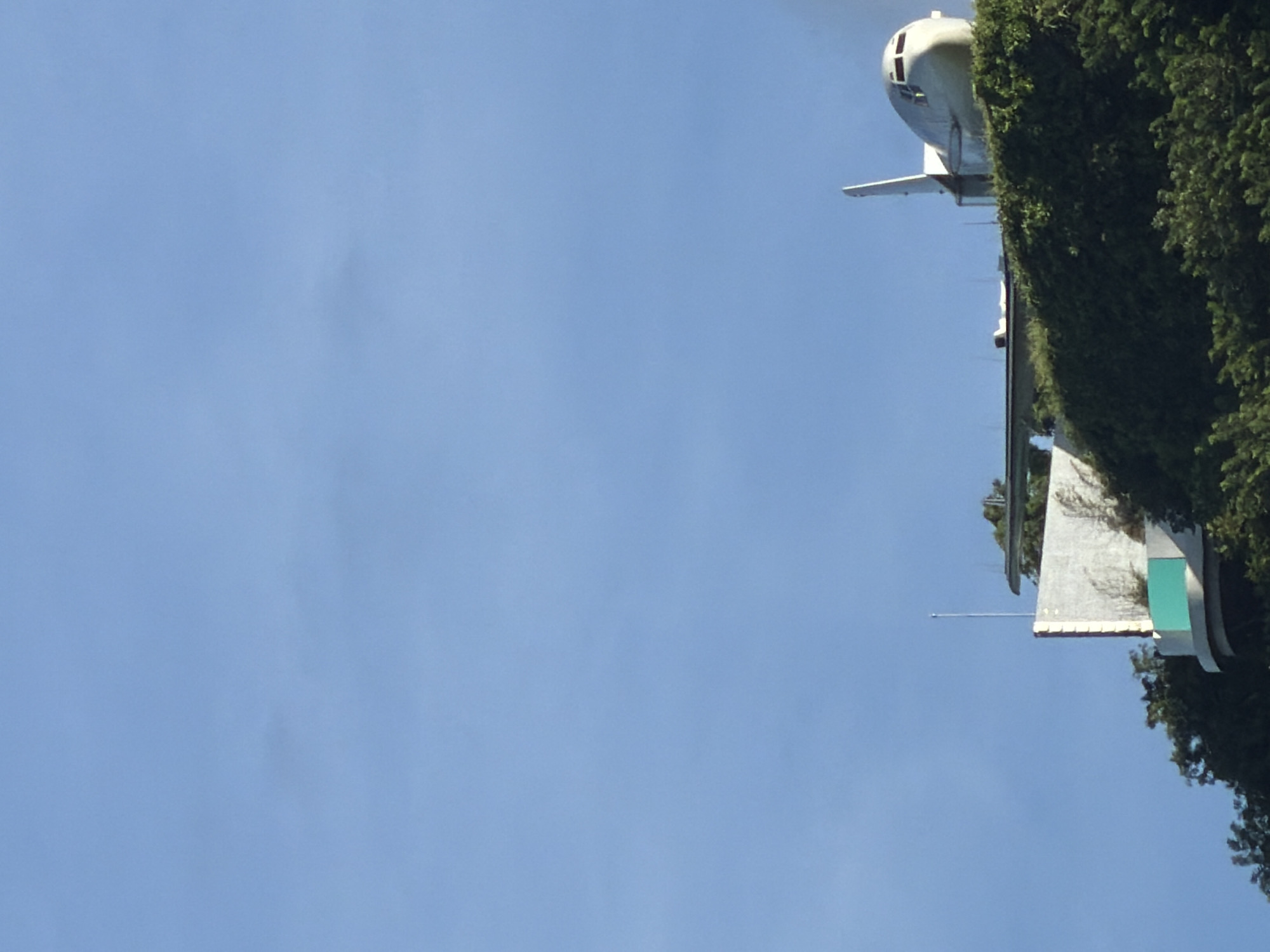 The image shows a view in Kuta Selatan, Indonesia, looking up at the sky. A partial view of a building is visible in the upper-right quadrant, partially obscured by green foliage. Above the building, a fuselage of what appears to be a decommissioned airplane is visible, its silver exterior contrasting with the clear blue sky. A thin antenna-like structure extends to the left from the side of the building.