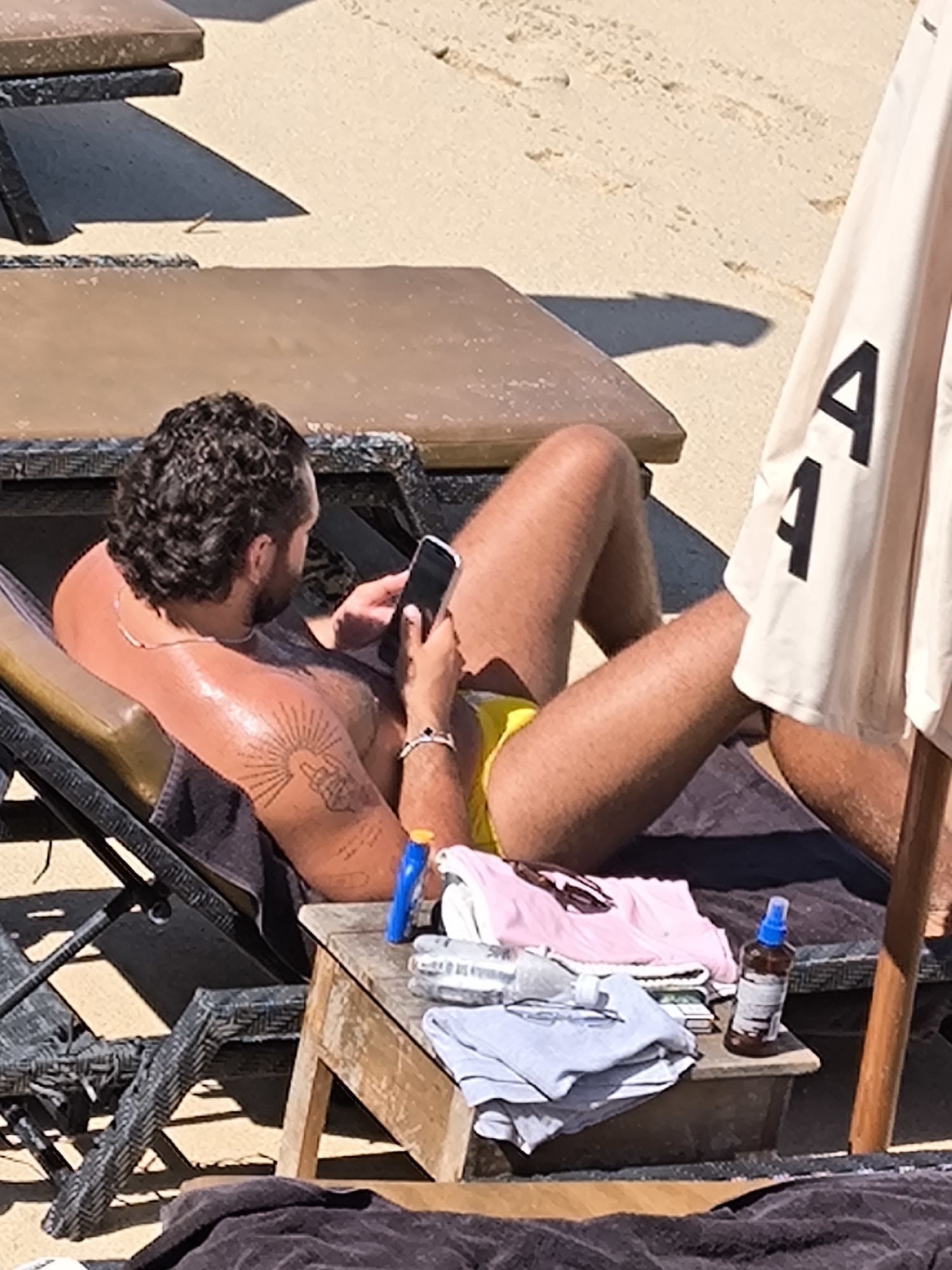 A man with curly dark hair, wearing yellow swim trunks, is relaxing on a lounge chair on a sandy beach. He is holding a smartphone in his right hand and appears to be looking at it. He has a tattoo on his left arm. Next to him on a small wooden table are folded clothes, sunglasses, a bottle of sunscreen, and a small spray bottle. A large umbrella with the letters "A" and "4" printed on it is partially visible to the right of the man. The scene is illuminated by bright sunlight, casting shadows on the sand. The overall environment suggests a beach resort or a public beach area in Kuta Selatan, Indonesia.