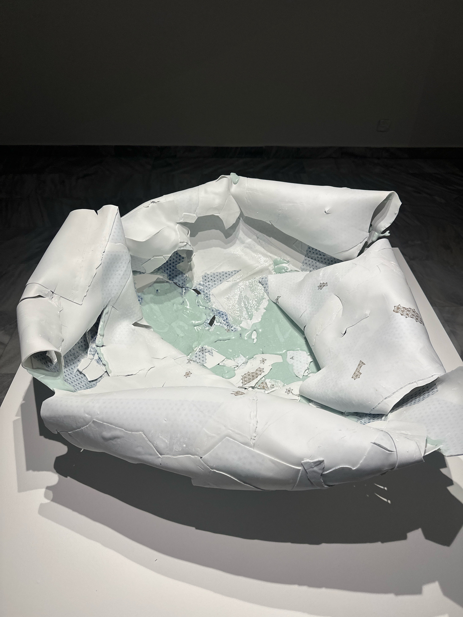 The image shows a white, fragmented sculpture with a patterned interior, presented on a white pedestal. The sculpture appears to be made of layered or peeling material, revealing a light blue and white geometric pattern beneath. The lighting is dim, casting shadows on the white surface and highlighting the texture of the sculpture. The background is a dark, indistinct wall and floor, suggesting an indoor exhibition space. The sculpture's location is not discernable from the image, and no context about Belgrade, Serbia is provided within the visual information.