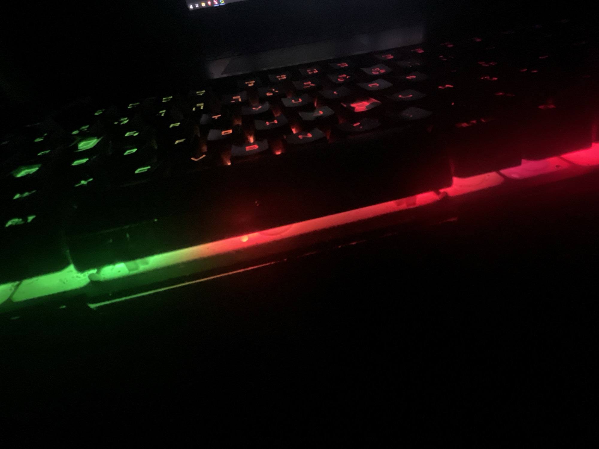 The image shows a close-up, low-angle shot of a gaming keyboard in a dark environment. The keyboard is illuminated by RGB lighting, with a gradient transition from green on the left side of the image to red on the right. The keys are black and appear to be backlit, with the light shining through the symbols on the keys. Some keys are partially obscured by the angle and the low light. The overall impression is one of a modern, technologically advanced setup. There is no indication of the city of Kołobrzeg, Poland, or any specific location within it. The scene is devoid of any people or discernible background details, focusing solely on the keyboard and its lighting effects.