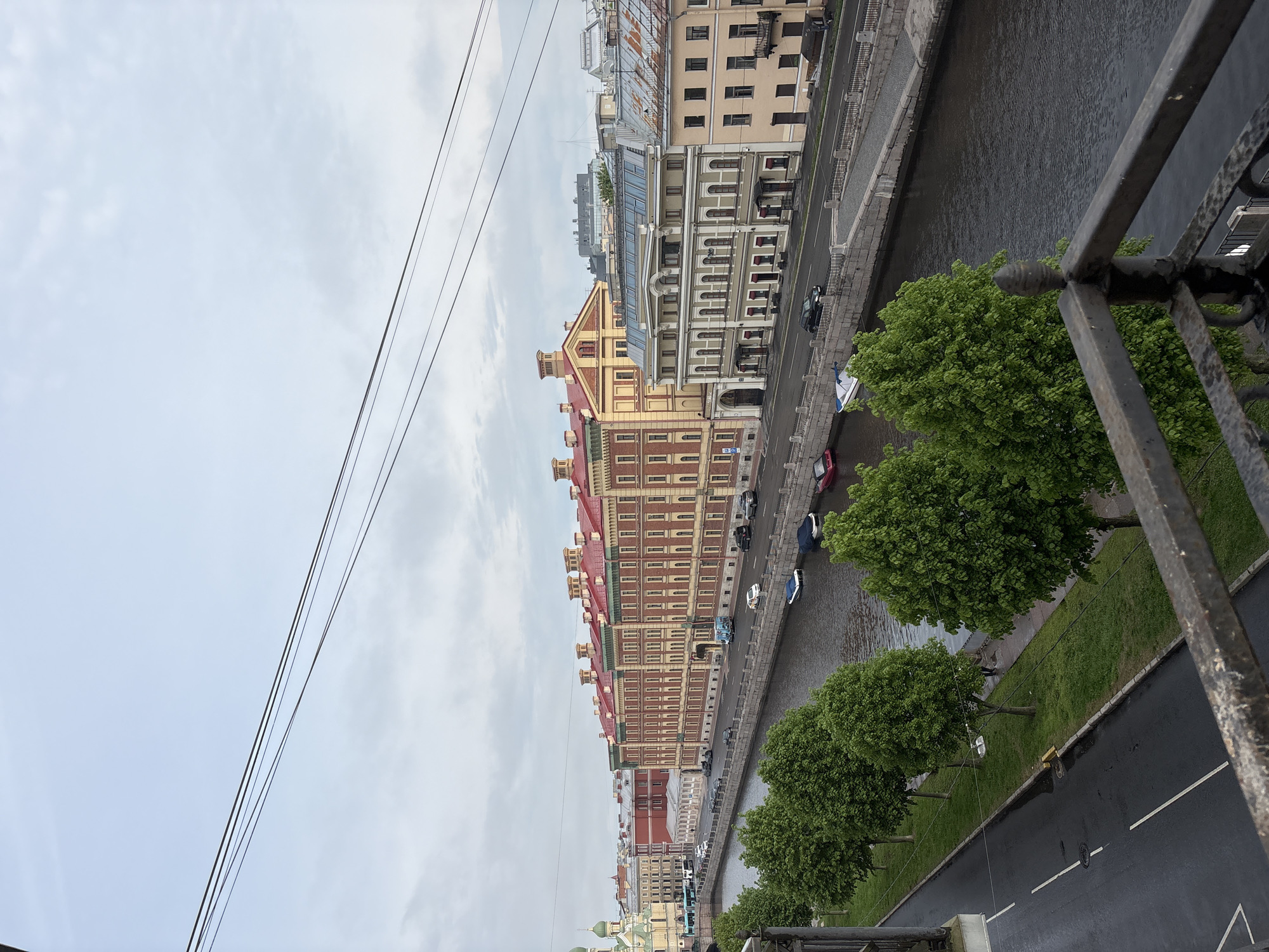 The image captures a scene along a canal in Saint Petersburg, Russia, viewed from an elevated perspective, likely a bridge. The buildings that line the canal have detailed architectural facades with varying colors and heights, featuring red and yellow hues. Below the buildings is a roadway adjacent to the canal. Cars are parked along the side of the roadway. The canal is bordered by a grassy area that has round green trees. The sky above is overcast. There are power lines visible in the upper-left corner. Part of a bridge railing is visible in the lower-right corner.