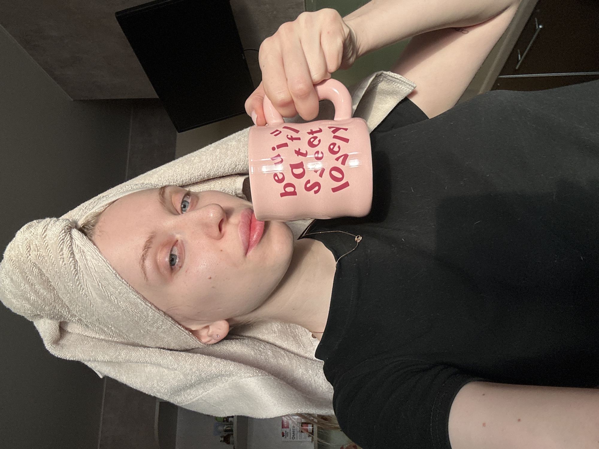 The image captures a fair-skinned individual, appearing to be a young adult female, engaged in a self-portrait within a domestic setting in the city of Chaikovsky, Russia. Her blonde hair is wrapped in a light beige towel. She wears a black short-sleeved t-shirt and a delicate gold-colored necklace with a small pendant. A small silver-colored stud earring is present in her left earlobe. She holds a textured, light pink ceramic mug with her right hand, positioned near her mouth. The mug features dark pink stylized text spelling "beautiful sweet lovely." A faint tattoo is partially visible on her right forearm. The background on the left displays shelves containing various items, including a box with "Омега" visible in Cyrillic. To her right, a dark flat-screen television or monitor is mounted on a dark grey wall, with a dark-colored cabinet featuring a silver handle situated below it. The scene is illuminated by natural light from the left side.