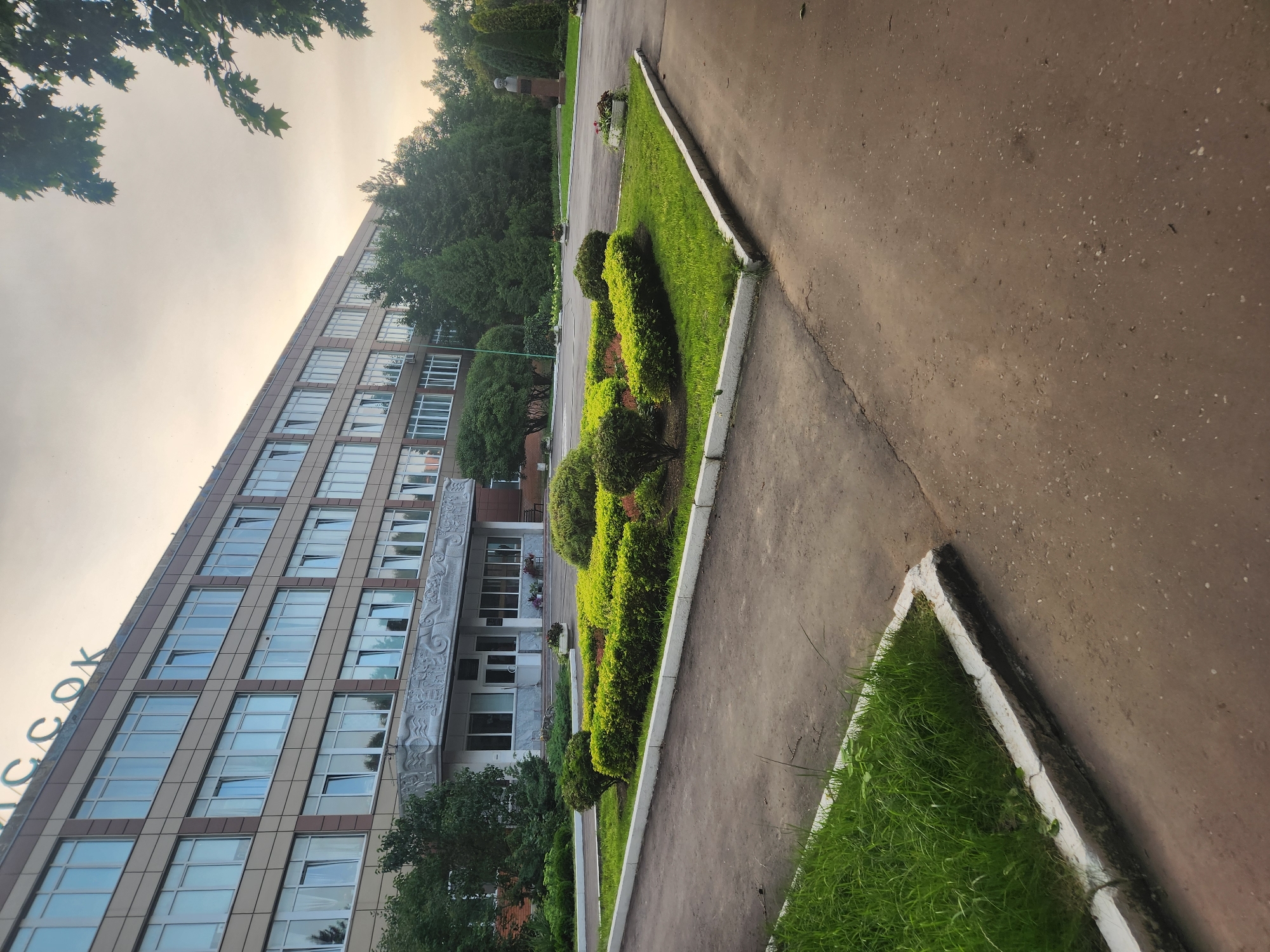 The image depicts the exterior of a large, multi-story building with a sign that reads "VNIISSOK" in green letters at the top. The building has a facade of beige panels and numerous windows with white frames. In front of the building, there is a well-maintained flower bed with manicured green bushes and some red flowering plants. A paved walkway or road curves around the flower bed, and to the left, there is a strip of green grass. The background features a line of trees and a cloudy sky. To the right, in the distance, a bust on a pedestal is visible. The overall scene suggests an institutional or corporate environment within Vniissok, Russia. There are no people visible in the image.
