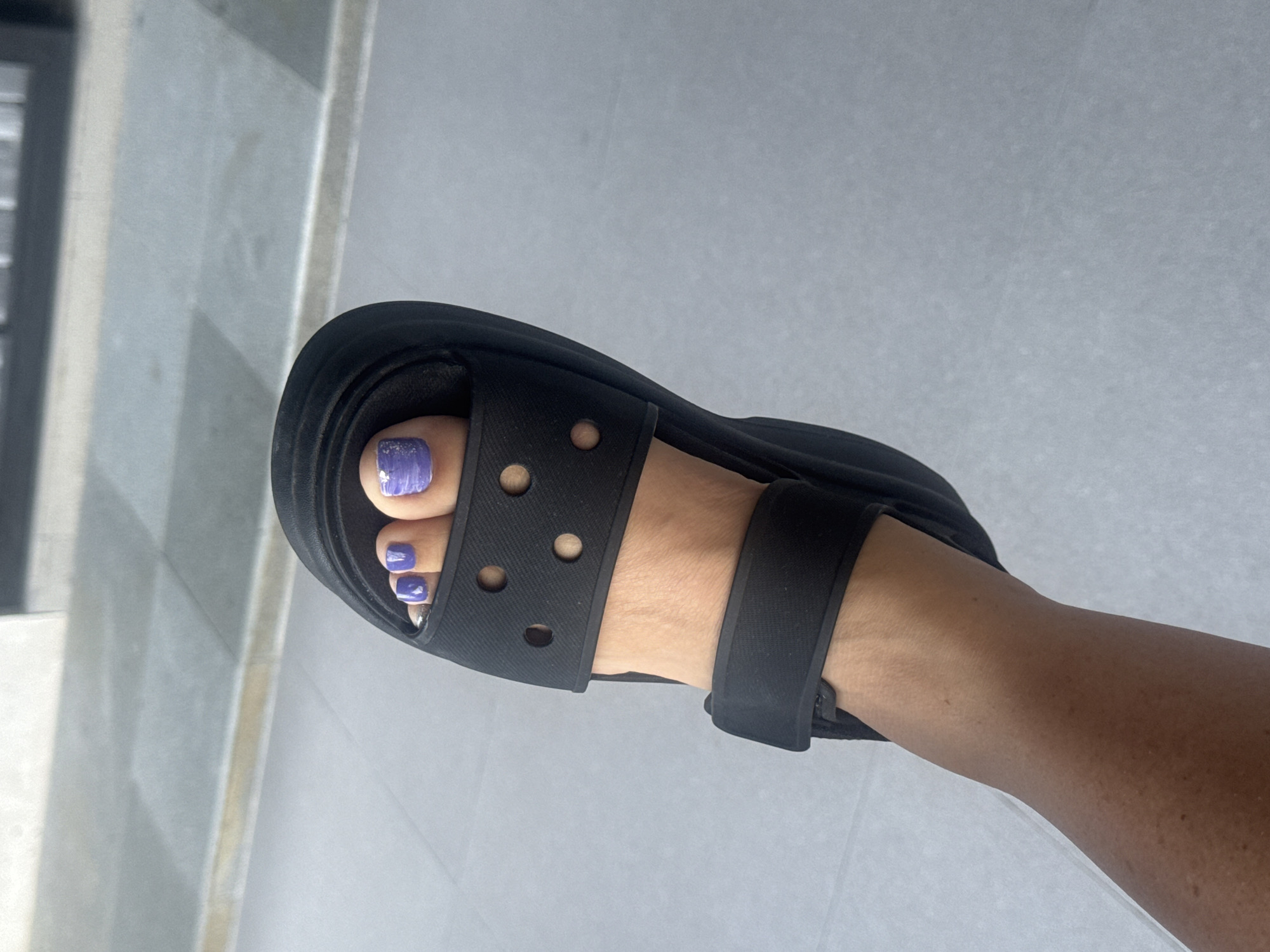 A close-up, overhead shot shows a person's left foot wearing a black platform sandal. The sandal has two straps, one across the toes and one around the ankle. The toes are painted with a lavender polish, with a touch of glitter on the big toe. The foot is resting on a light gray tiled floor, with a blurry background that appears to be part of an outdoor public space.