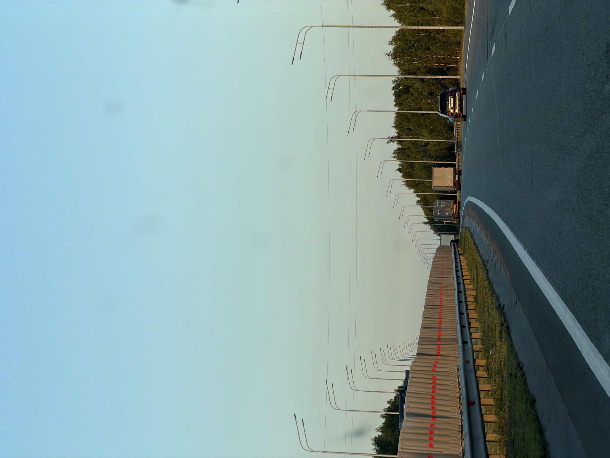 The image shows a view from a car traveling on a highway. The sky is clear and a light blue, with a hint of yellow at the horizon. A line of concrete barriers with red reflective markers runs along the left side of the road. Beyond the barriers, several streetlights are visible, lining the highway. A dense line of green trees forms the background on the right side of the image. Two trucks are visible ahead, traveling in the same direction as the car, followed by a dark SUV. The road is asphalt with white lane markings.