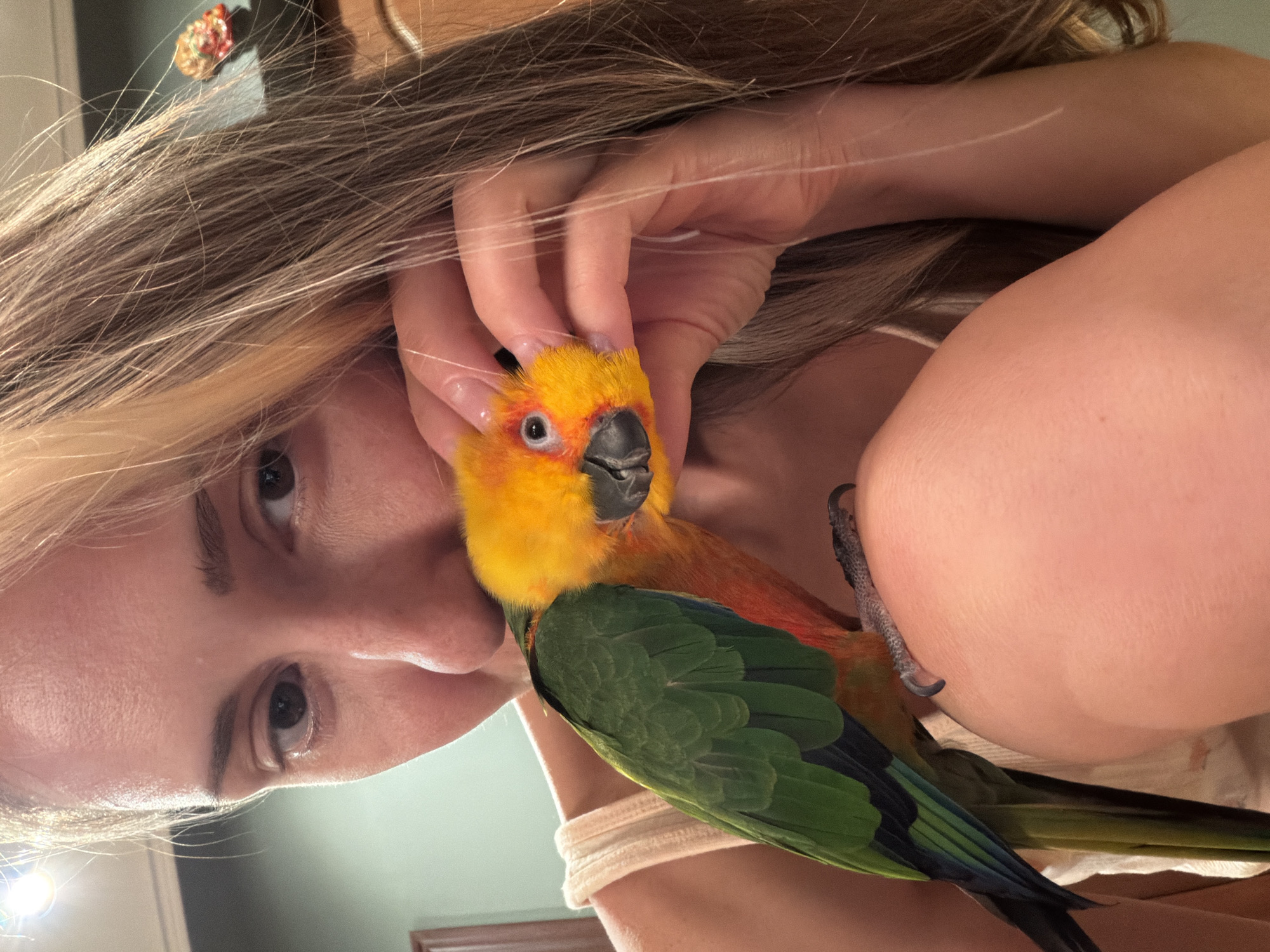 A woman is holding a Sun Conure parrot. The woman has fair skin, brown eyes, and blonde and brown hair. She is gently holding the bird's head with her fingers. The parrot is brightly colored with a yellow head, orange chest, and green wings. The background is blurry and indistinct. There is no visible indication of the city of Khimki, Russia.