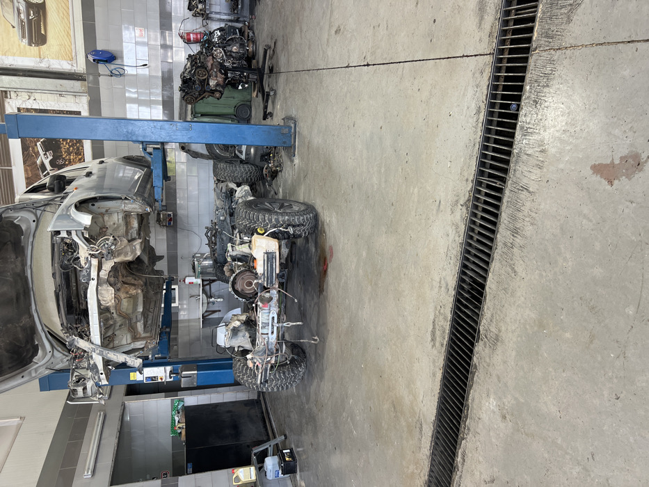 A car body is lifted by a blue hydraulic lift in a garage setting. Beside it, the car's chassis with wheels is present, suggesting disassembly. Scattered car parts and tools fill the background, and a floor drain runs along the edge of the concrete floor, indicating a repair facility. The overall scene suggests a complex automotive repair or restoration project is underway.
