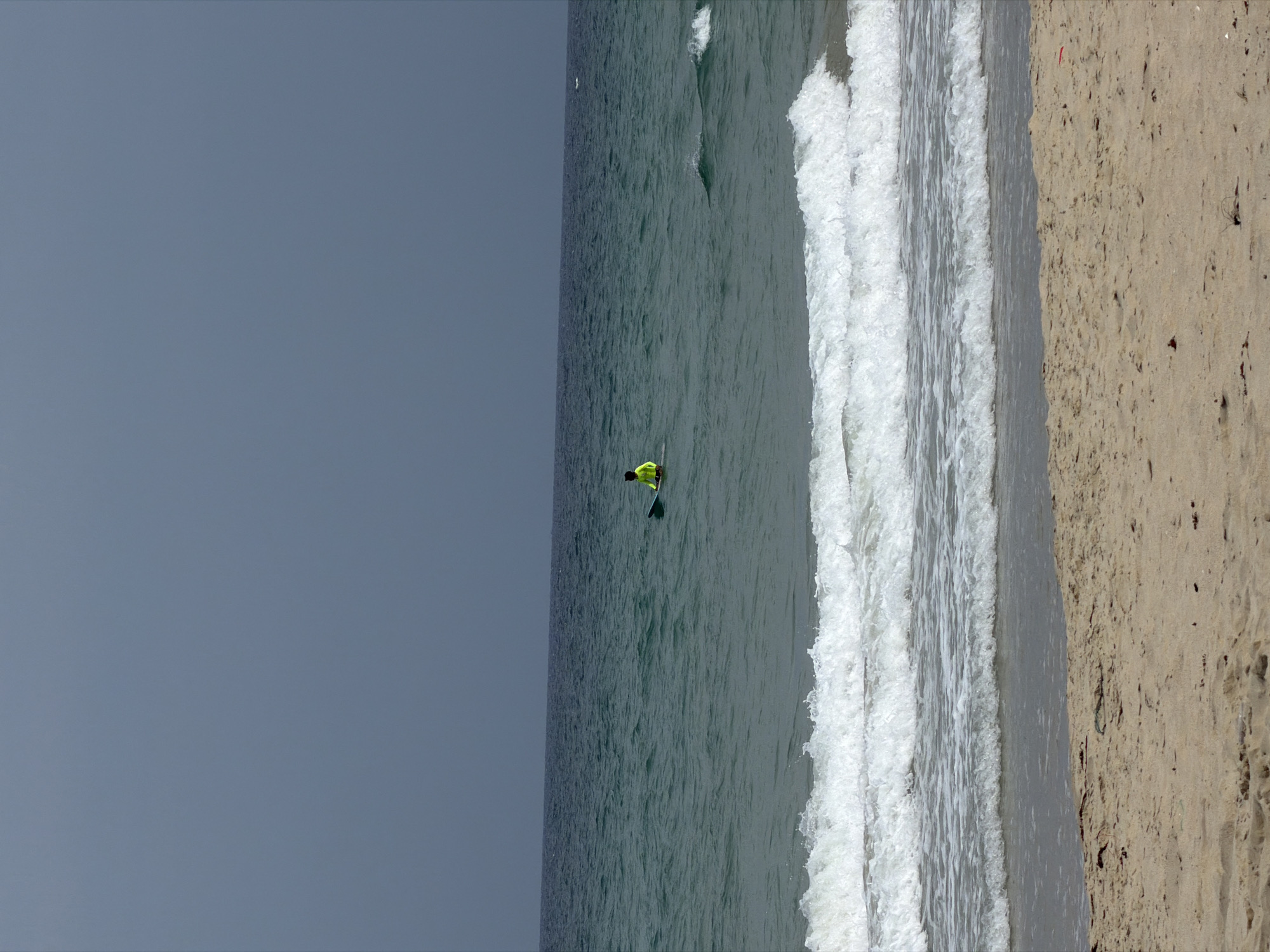 A sideways view of a seascape featuring a surfer in the city of Cam Lâm, Vietnam. A body of water stretches across the middle of the frame, with waves breaking near the coastline. A person, dressed in a bright yellow garment, stands on a paddleboard in the water, accompanied by a paddle. The left of the frame consists of a solid gray expanse suggesting the sky or empty space. To the right of the water, there's a portion of sandy beach.