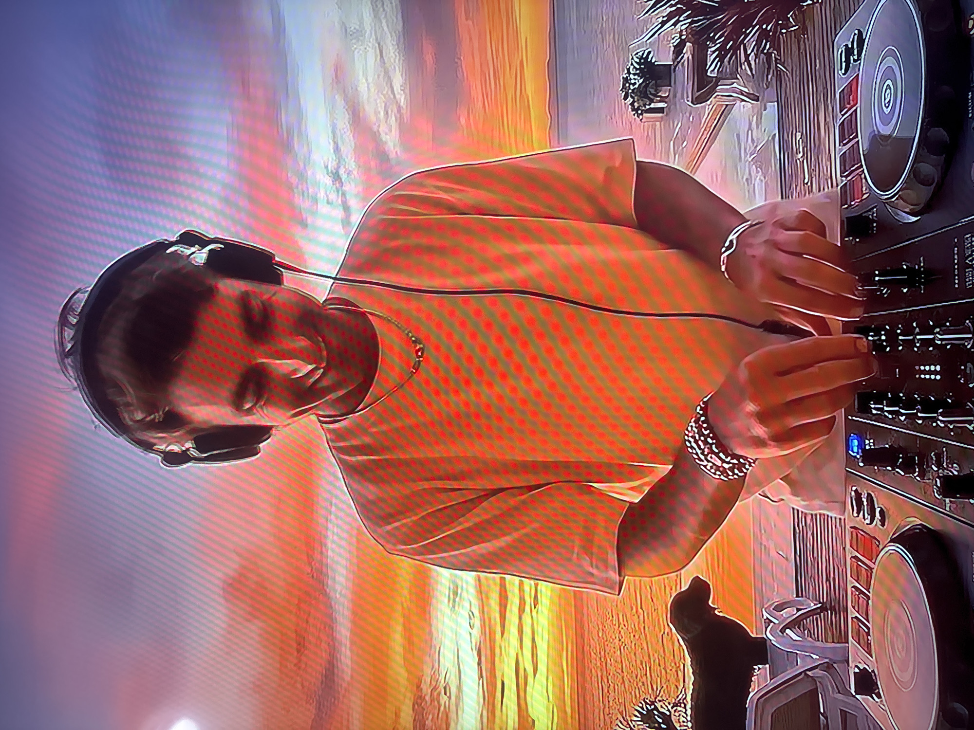 The image shows a young man wearing headphones and a t-shirt, standing behind a DJ setup. He is smiling and appears to be mixing music. The background depicts a sunset over a body of water, with palm trees and lounge chairs visible. A dog is sitting on a chair in the left side of the image. The man is wearing a necklace and bracelets. The DJ setup includes two turntables and a mixer. The overall scene suggests a relaxed and festive atmosphere, possibly a beach party or a resort setting. There is no visual information to suggest the location is the city of Krāslava, Latvia.