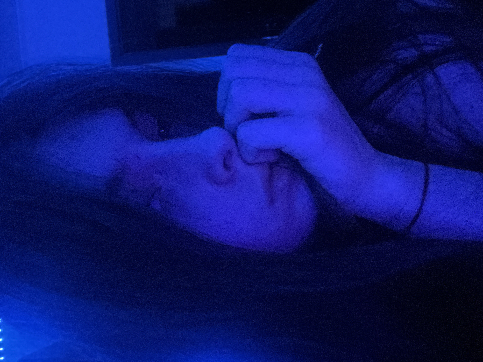 The image displays a person, likely a young female, illuminated by a dominant blue light source originating from the upper left quadrant. The individual is positioned facing towards the right side of the frame, with their head slightly tilted downwards and eyes cast away from the viewer. Their right hand is resting with fingers curled against their chin and lower lip, suggesting a pensive or contemplative state. Long, dark hair frames their face and extends over their shoulders. A thin, dark band is visible on their right wrist. The background is largely indistinct due to the low blue illumination and deep shadows. In the upper left corner, several small, bright points of light, consistent with an LED strip, are visible. Faint architectural lines, possibly a doorframe or wall edge, can be discerned on the right side of the frame. The scene appears to be indoors. The image exhibits noticeable digital noise or grain, characteristic of low-light photography. No identifiable landmarks, signage, or other contextual clues are present within the image to indicate a specific geographic location, including the city of Saint Petersburg, Russia.