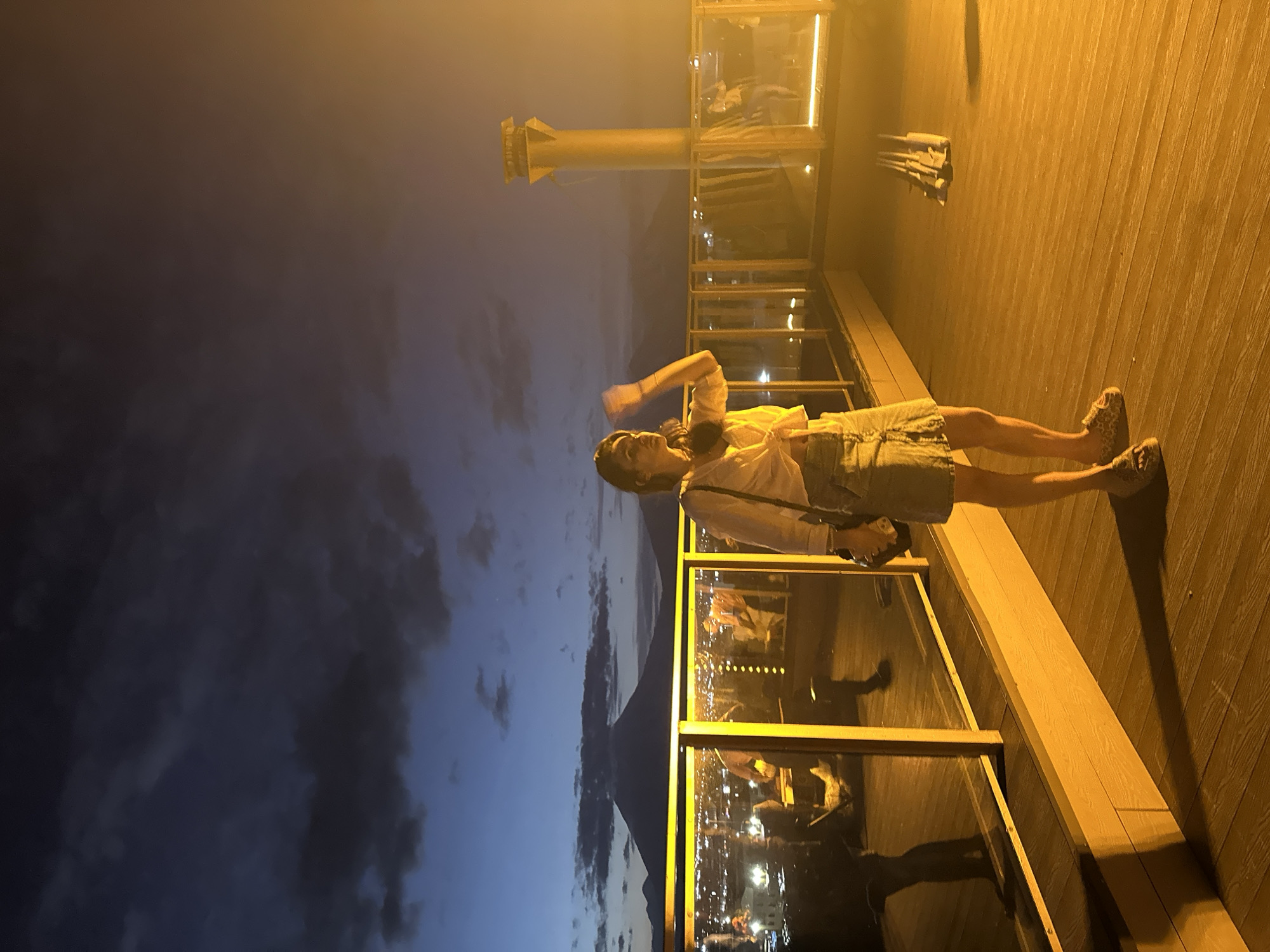 The image depicts a nighttime scene from a rooftop in the city of Petropavlovsk-Kamchatsky, Russia. In the foreground, a young woman is standing on a wooden deck, illuminated by artificial lights. She is wearing a white, knotted shirt and a denim skirt, with leopard-print sandals. She is holding a dark bag on her shoulder and appears to be raising her right hand. Behind her, a glass railing separates the deck from a lower level where city lights are visible, suggesting a bustling urban environment. In the background, a large, dark silhouette of a mountain is prominent against the twilight sky, characteristic of the volcanic landscape surrounding Petropavlovsk-Kamchatsky. The sky is a deep blue, transitioning to darker hues with hints of clouds. To the right, a tall, cylindrical structure, possibly a ventilation shaft or part of the building's architecture, is brightly lit with a warm, orange glow. The overall atmosphere is one of a serene evening with a panoramic view of the city and its natural surroundings.