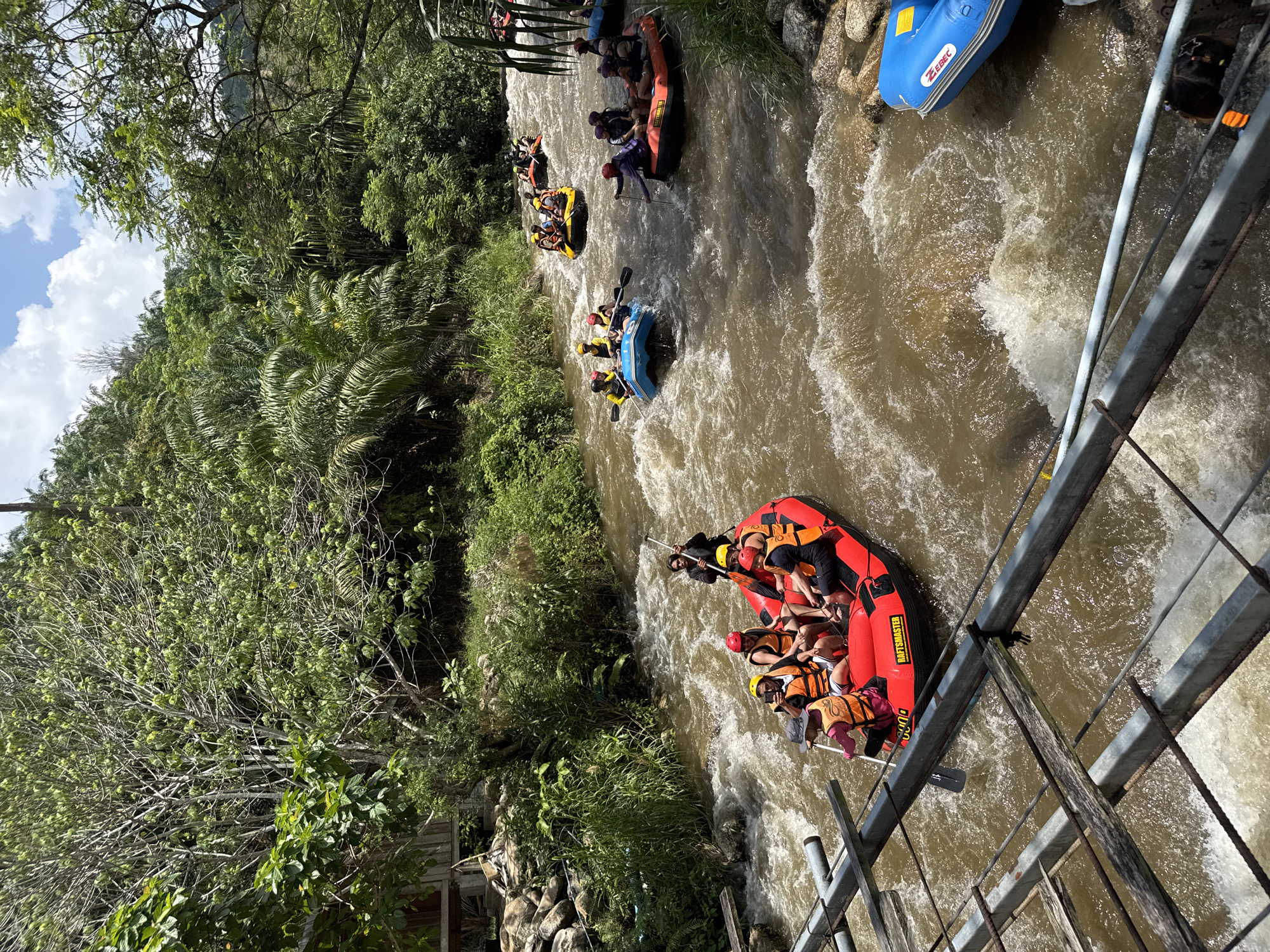 The image shows a whitewater rafting scene on a river in Tambon Song Phraek, Thailand. Multiple inflatable rafts filled with people are navigating through rapids. The rafts are of different colors including red, blue, yellow, and black. The people in the rafts are wearing life jackets and using paddles to steer. The river is surrounded by lush green vegetation, including trees and bushes. A metal bridge railing is visible in the foreground. The water is choppy and brown, indicating fast-flowing currents.