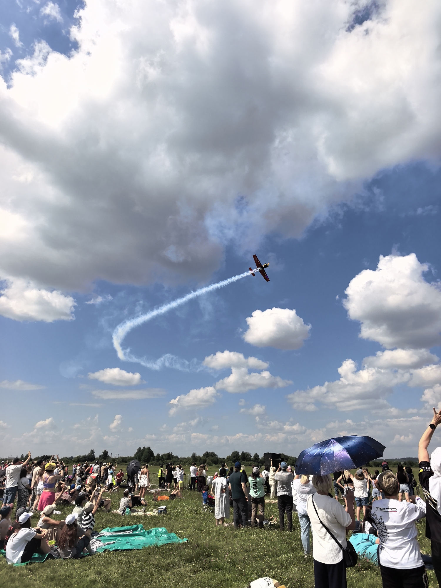 The image depicts an aerial acrobatic performance with a small red and yellow airplane trailing a white smoke stream against a blue sky filled with white and grey clouds. The plane is viewed from below and slightly to the side, mid-maneuver. Below the sky, a large crowd of people are gathered in a grassy field, many sitting on blankets and chairs, observing the event. Some individuals are standing, and a few are captured with their arms raised. The environment appears to be an outdoor event, possibly an airshow. There is no visible indication of the city of Perm, Russia, within the image itself.