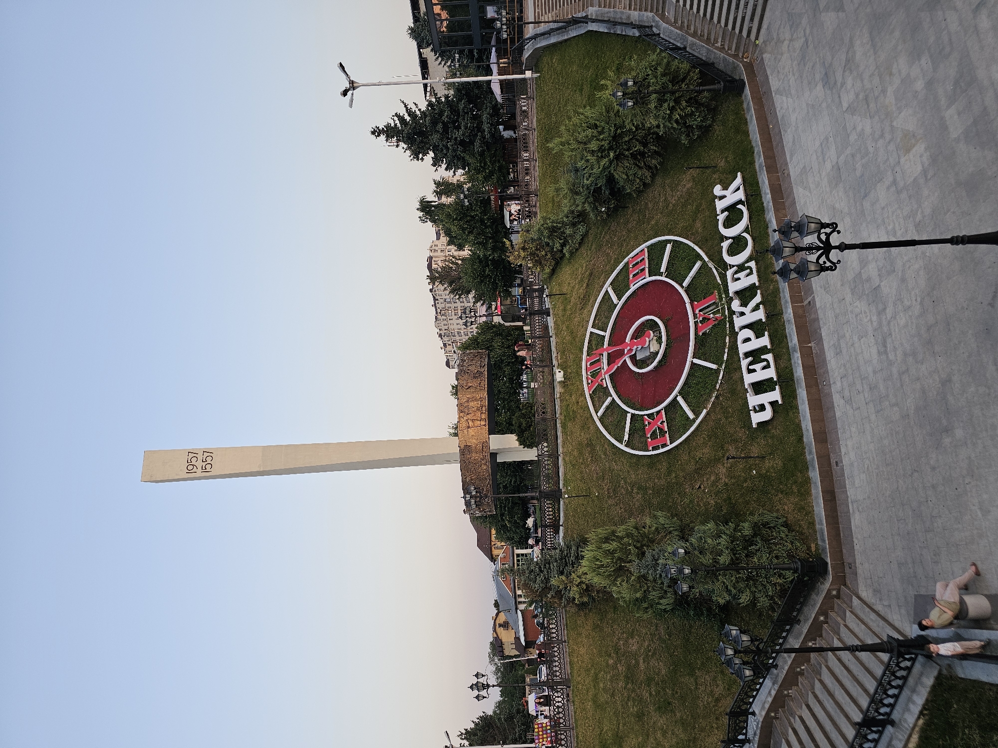 The image depicts a public square in Cherkessk, Russia, featuring a large obelisk monument and a decorative clock on a grassy embankment. The obelisk, a tall, slender structure, has the years "1957" and "1557" inscribed on it. At the base of the obelisk is a circular frieze. Below this, on the sloping lawn, a large clock face is prominently displayed, with Roman numerals and red clock hands. The words "ЧЕРКЕССК" (Cherkessk) are spelled out in large white letters on the embankment below the clock.
The environment includes a paved plaza area with cobblestones, interspersed with lampposts. A set of wide stone stairs leads up to the plaza. On the plaza, two individuals are seated and standing near a bench. Several trees, including evergreens, are planted on the embankment and in the background. Buildings, some with multiple stories, are visible in the distance behind the monument and trees. The sky is clear and light blue, suggesting it is daytime. The scene appears to be a public gathering space or monument area in the city.