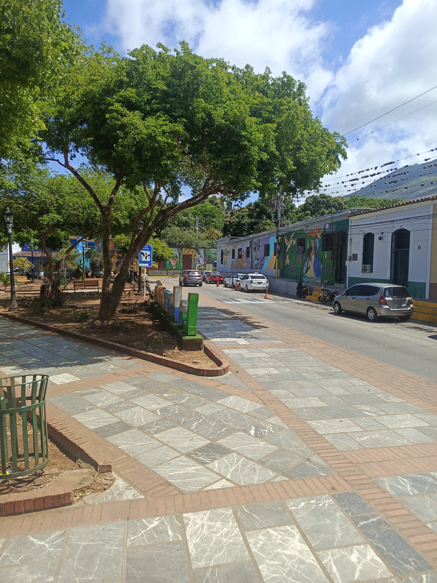 The image depicts a street scene in La Asuncion, Venezuela. A paved plaza, constructed of grey and brick tiles, dominates the foreground. A green metal trash can stands on the left. A lush green tree provides shade to a park area on the left, which includes benches.
Across the street, parked cars are visible, including a silver hatchback and a white taxi. Buildings with colorful painted murals line the street. A mountain rises in the background. The sky is blue with scattered clouds. A street sign is visible near the park area.