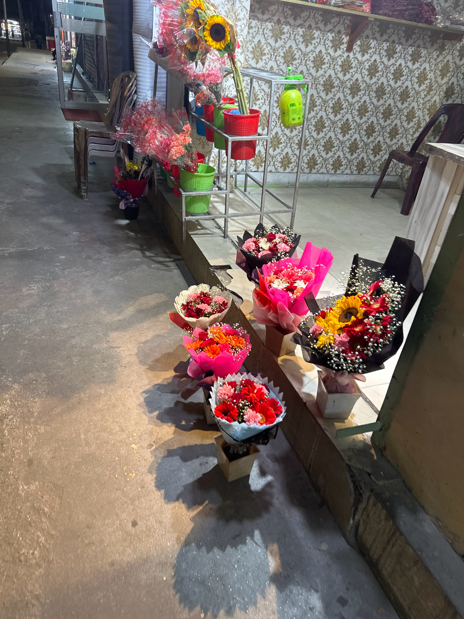 A flower stall is set up on the side of a street in Kathmandu Metropolitan City, Nepal. The stall features a metal rack holding several buckets of flowers, including sunflowers and carnations, in various colors such as red, pink, and yellow. Some bouquets are wrapped in black and pink paper, while others are in white paper. A green spray bottle hangs from the rack. The ground is paved with concrete. In the background, a shop with patterned wallpaper and a dark brown chair can be seen. Several stacks of chairs are placed in front of a building. The lighting suggests it is nighttime.