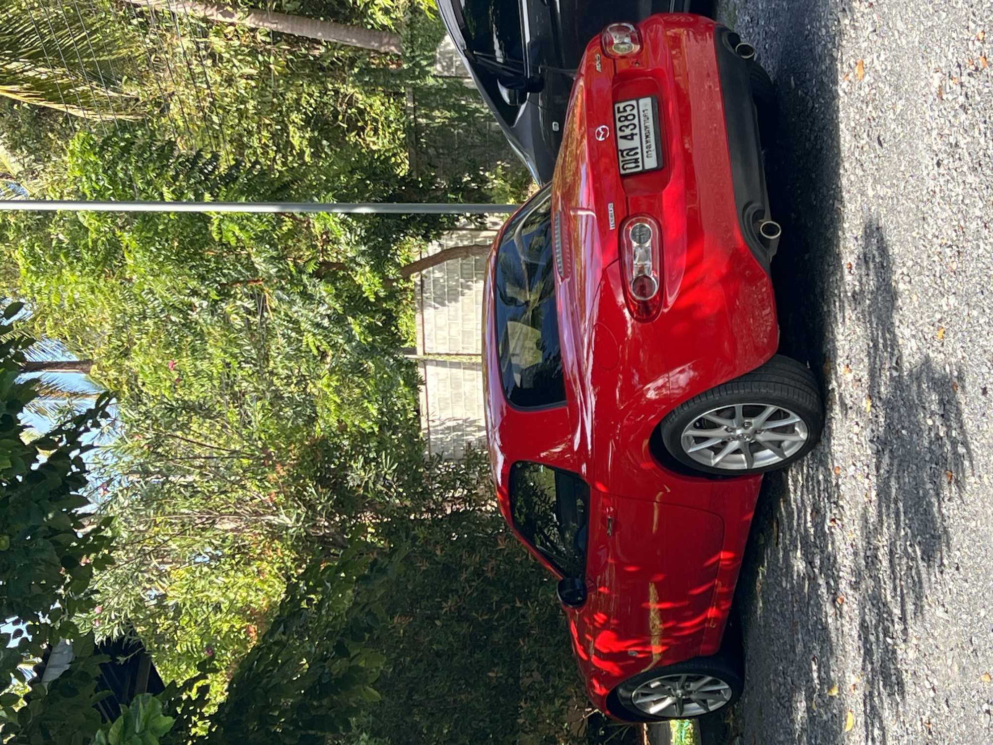 In บ้านบางรักษ์, Thailand, a red Mazda MX-5 Miata with a hardtop is parked on a gravel surface, casting a shadow from overhead foliage. The car's rear license plate reads "กส 4385". Beside it, the rear of a dark-colored vehicle is visible. The background is dominated by lush green trees and foliage, with a hint of a concrete wall and a street lamp. The lighting suggests it is daytime, with dappled sunlight filtering through the leaves.