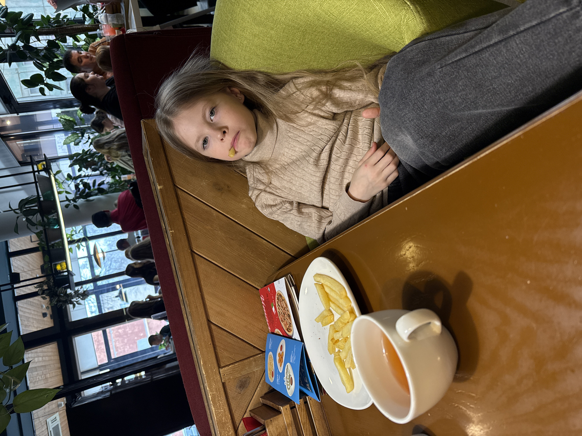A young, blonde girl is lying on a lime-green, upholstered bench seat at a restaurant. She has a french fry in her mouth and is wearing a beige turtleneck sweater and gray pants. On the wooden table in front of her are a white plate with french fries, a menu, and a white cup of tea. In the background, other people can be seen sitting at tables and looking out the window. Through the window, the outside environment is visible. The location is the Mesto cafe in Izhevsk, Russia.