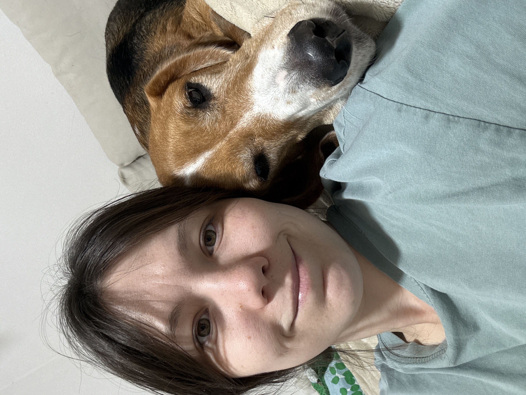 A woman is lying on a surface with a beagle dog lying on her chest. The woman has brown hair and hazel eyes. She is wearing a light blue-green shirt. The dog has a black nose and brown and white fur. The background is a white wall. The location is not discernible from the image.