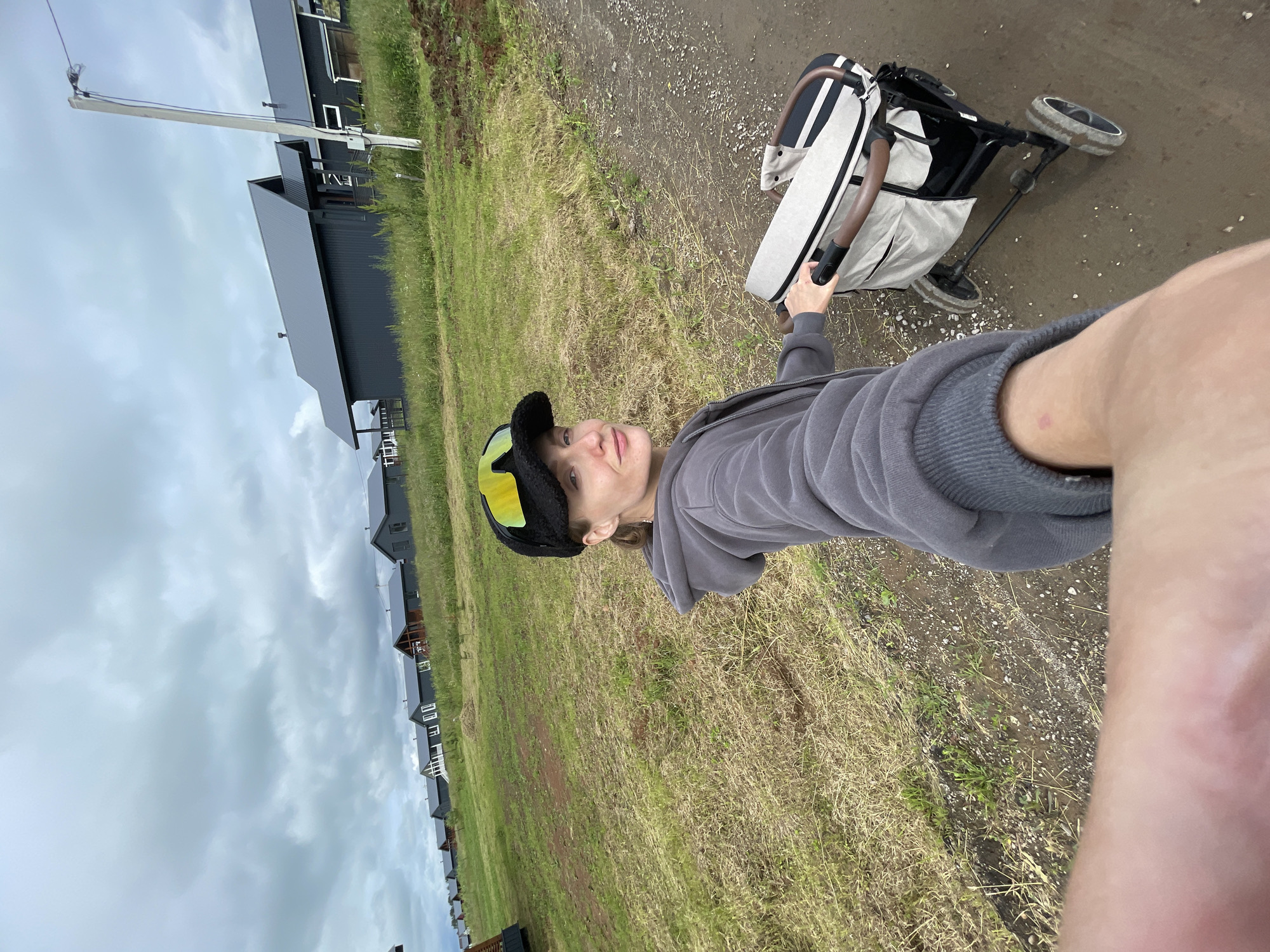 A person, wearing a grey hoodie and a black baseball cap with yellow and black goggles, is captured in a selfie. They are pushing a stroller with a beige and striped bag. The stroller has dark wheels and brown handles. The person's right arm and leg are partially visible in the foreground. The scene appears to be outdoors, with a path or dirt road beneath the person and stroller, bordered by dry grass and some greenery. In the background, a row of dark, modern-style houses with dark roofs are visible, situated on a slight incline. The sky is overcast and cloudy. The overall mood is casual and possibly reflective. The time of day is not explicitly indicated but appears to be daytime given the light. The location is identified as Podshivalovo, Russia. No visible text is present in the image.