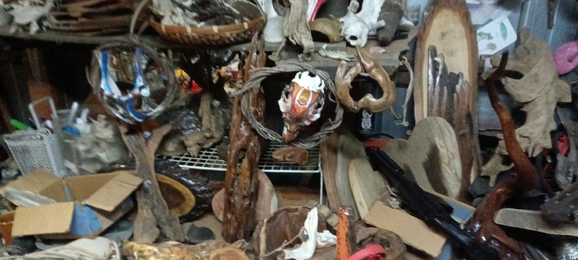 The image appears to be a cluttered indoor space, possibly a storage area or workshop, located in Tahlequah, United States. The scene is dominated by various objects including driftwood, woven baskets, and what appears to be animal skulls or bones. These items are arranged on shelves and the floor, giving the impression of disorganization and storage. There are no people visible in the image. The environment is dimly lit, contributing to the cluttered and haphazard appearance. The focus seems to be on the collection of natural and possibly crafted objects, suggesting a connection to nature, art, or perhaps a collection.