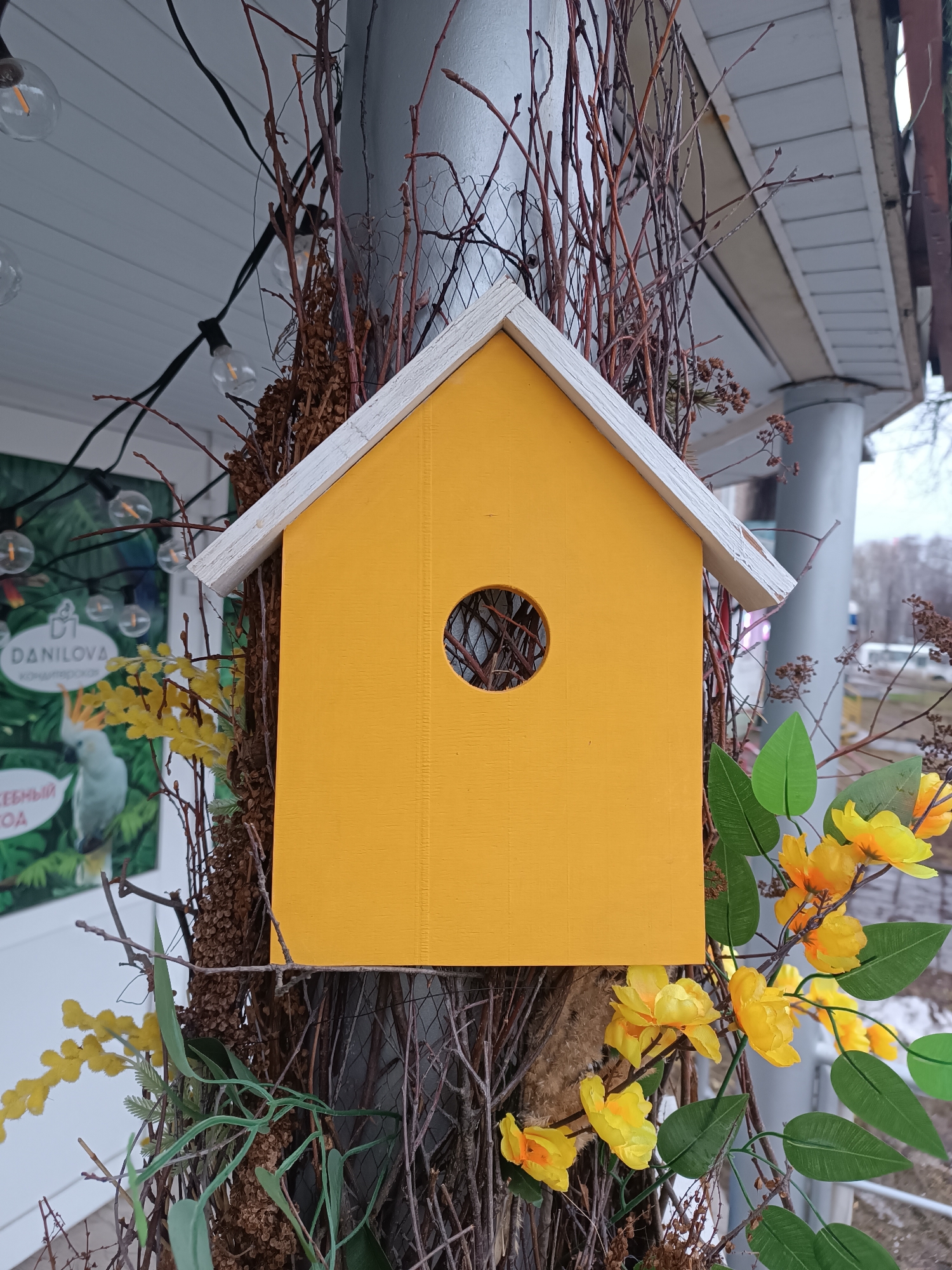 A yellow birdhouse with a white roof is mounted on a gray metal pole, surrounded by dried branches and artificial yellow flowers with green leaves. The birdhouse has a round opening. In the background, a sign with a bird and the name "DANILOVA" in Russian is visible, along with decorative light bulbs. The scene appears to be in an urban setting, specifically Chaikovsky, Russia. A street with some vehicles and bare trees can be seen in the blurred background.