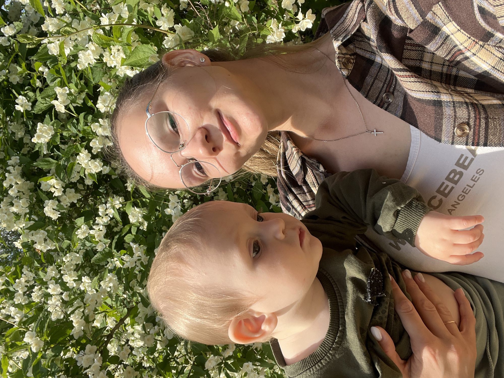 An outdoor, rotated close-up portrait shows a young woman holding an infant against a backdrop of dense, white flowering jasmine bushes in Chaikovsky, Russia. The woman, positioned in the upper left, has light hair tied back and wears round-framed glasses. She looks directly at the camera with a composed expression. Her attire includes a brown and white plaid shirt over a white tank top, and a silver cross pendant necklace is visible.
The infant, held by the woman, is in the lower right section of the frame, gazing slightly to the right with a curious expression. The baby has light hair and wears an olive green long-sleeved top. The woman's hand, adorned with a simple ring and light-colored nails, rests gently on the baby's chest.
The scene is brightly lit by strong sunlight, indicating a clear day. The background is a vibrant tapestry of green leaves and numerous small white flowers, typical of jasmine in full bloom during spring or early summer. Visible text on the woman's white tank top reads "CEBE ANGELES". The image conveys a serene and tender moment between the subjects surrounded by nature.