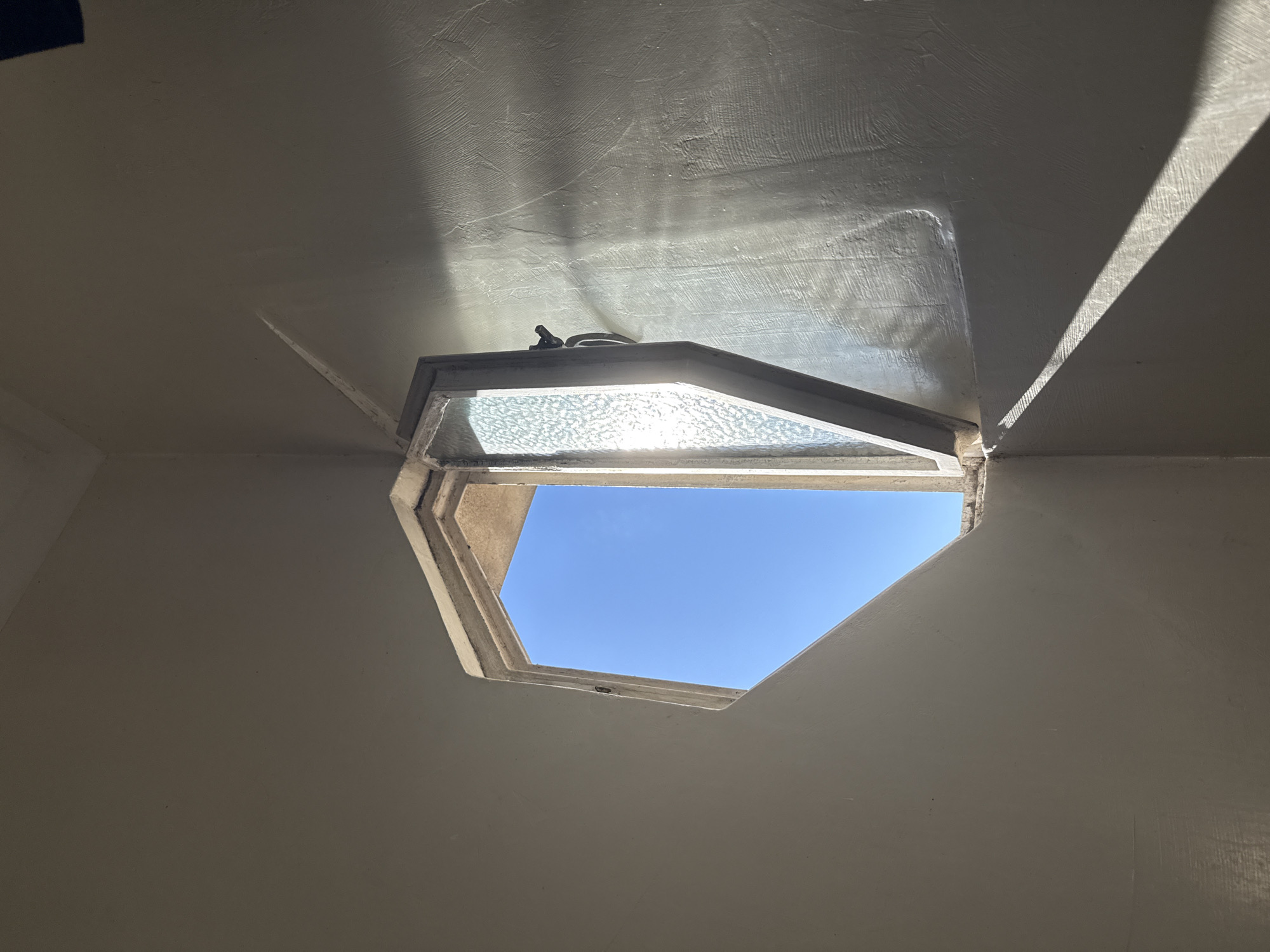 The image captures an upward-looking view of a distinctive ceiling skylight or window, showcasing a bright, clear blue sky. The window, appearing to be hexagonal or octagonal in shape, is partially open, with the bottom section directly exposing the sky. The upper section of the window frame contains a textured glass pane, through which bright sunlight streams, casting reflections and patterns onto the white or light-colored ceiling. A small black latch mechanism is visible on the upper frame of the window.
The ceiling itself is painted in a light, neutral color, possibly white or off-white, and exhibits a textured finish, with visible brushstrokes or plasterwork. Strong contrasts of light and shadow are evident on the ceiling, indicating bright daytime lighting. Direct sunlight creates defined streaks of shadow on the right side of the ceiling and illuminates the textured surface near the skylight, highlighting its uneven finish.
The visible area outside the window is solely a vibrant, cloudless blue sky, suggesting fair weather. No specific activity is depicted, but the scene emphasizes natural light and architectural design. There is no visible text. The setting is an interior space, likely in Buenos Aires, Argentina, offering a serene view of the sky above.
