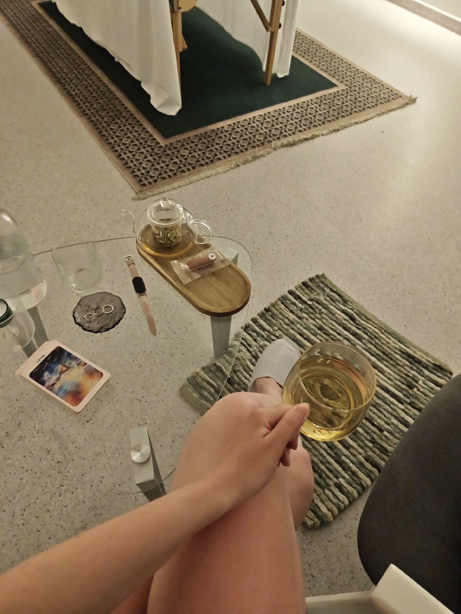 A high-angle shot reveals a person seated in a chair, holding a glass of liquid, likely wine, while resting their legs on a small, striped rug. They are wearing a grey slipper on one foot. Beside them is a glass-topped table with various items: a tarot card, a water bottle, a glass, rings on a stone coaster, a smartwatch, and a teapot with a wooden tray. Behind the table is a larger rug, with a massage table partially visible on top. The environment suggests a relaxation or wellness setting, possibly a private residence. The image does not provide enough information to determine if the location is in Vinnytsia, Ukraine.