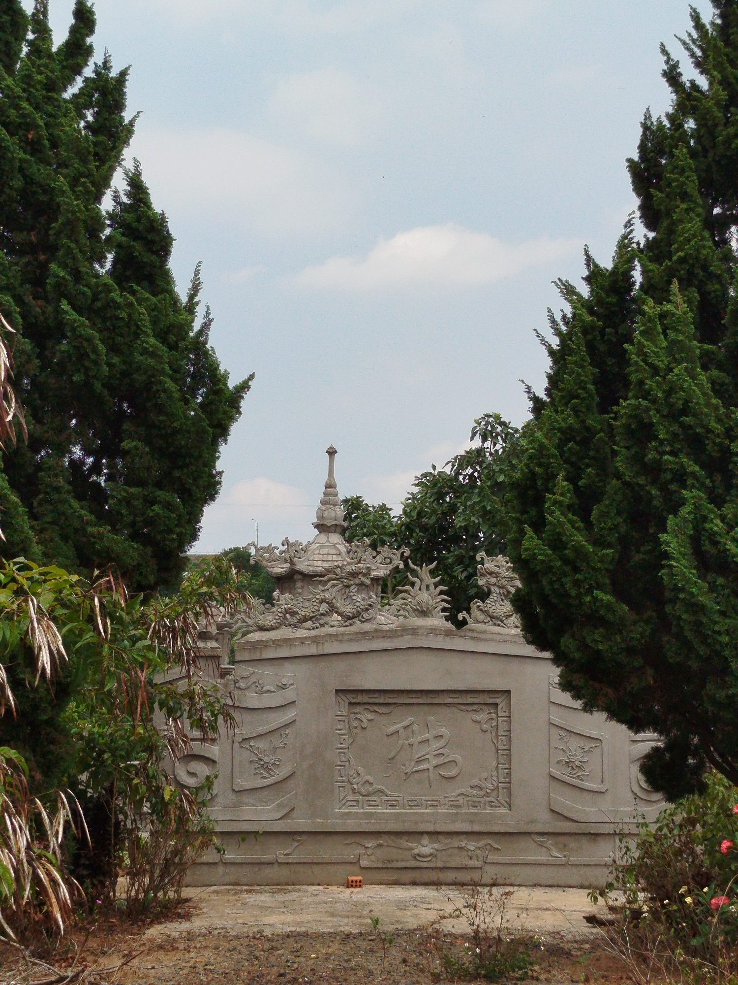 The image shows a stone wall with intricate carvings and a pagoda-like structure on top, framed by two tall, green cypress trees. The wall features a large rectangular panel with carved characters, possibly in Chinese or Vietnamese. Above the panel, there are sculpted dragons and other ornate decorations. The ground in front of the wall is bare earth with some small plants. A small, red brick is visible at the base of the wall. The sky above is light blue with scattered white clouds. The location is implied to be in the city of Liên Nghĩa, Vietnam. There are no people visible in the image.