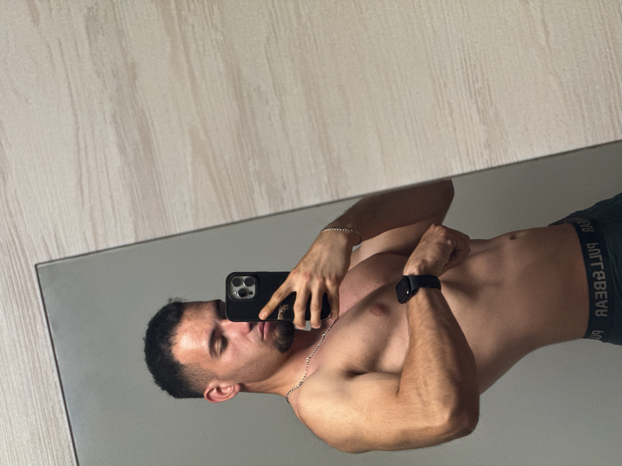 A man is taking a selfie in a mirror. He is shirtless and wearing dark colored underwear. He is also wearing a watch and a chain. The mirror is resting on a light wooden surface. The location in the city of Shumen, Bulgaria cannot be determined from the image.