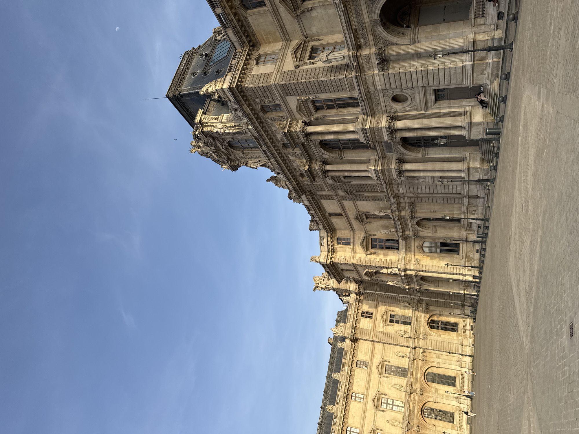 The image shows a view of the Louvre Palace in Paris, France, on a clear day with a blue sky. The building is a large, ornate structure with many windows, columns, and architectural details. The façade is made of light-colored stone. A portion of the pavement in front of the building is also visible. There are a few people on the steps in front of the building, and some light posts. In the sky, a tiny moon can be seen.
