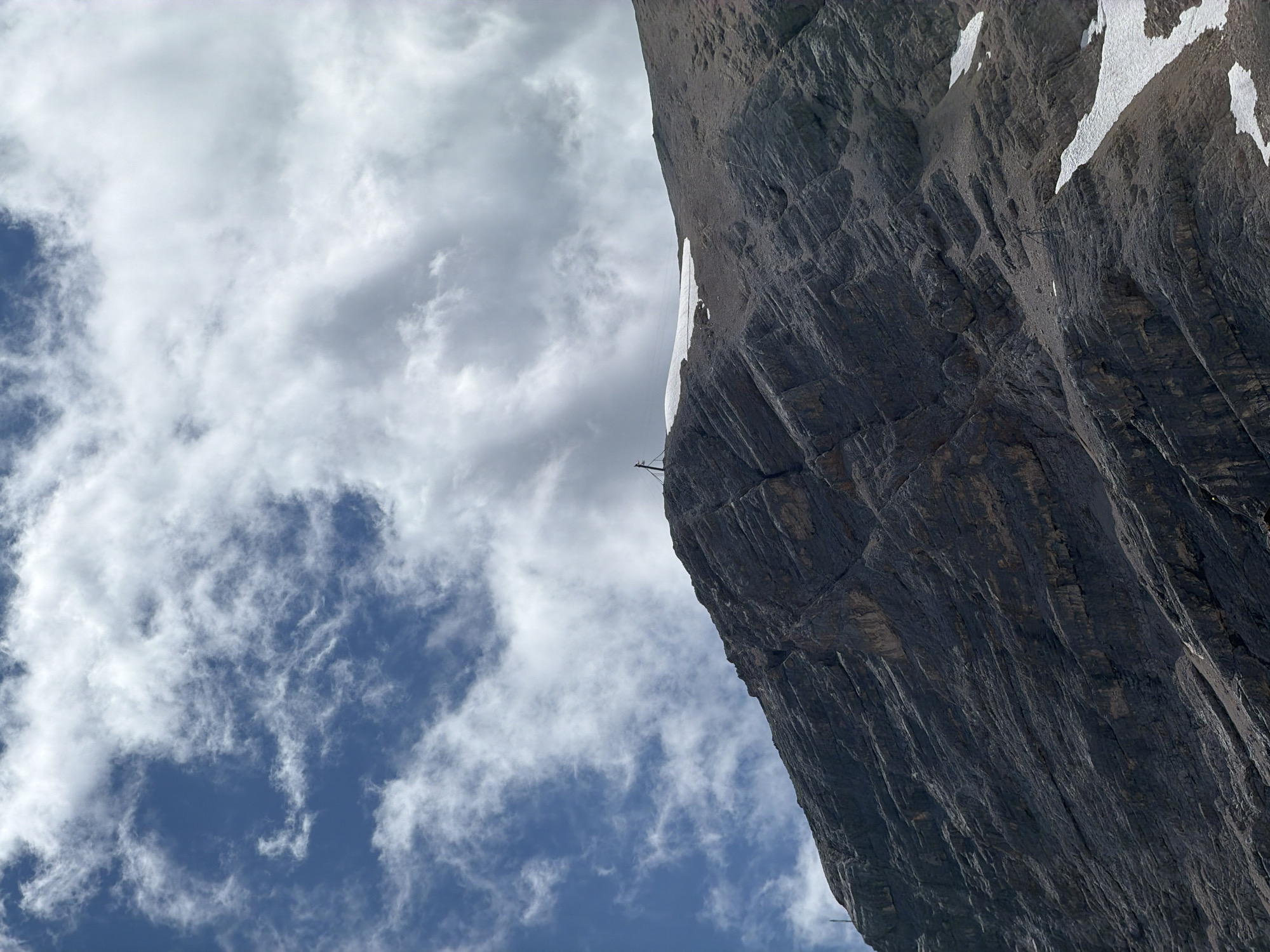 The image shows a close-up of a steep, rocky mountainside under a cloudy blue sky. The mountain is primarily composed of dark gray rock with visible stratification and texture, suggesting a rugged terrain. Patches of snow are present on the upper slopes, particularly on the right side of the image.
At the crest of the mountain, a small, dark, cross-shaped structure is visible, possibly a marker or monument. There are also some thin, dark lines extending upwards from the ground near the structure, which could be part of a support or antenna. The sky is filled with a mix of white and gray clouds, with patches of blue sky showing through.
While the image depicts a natural mountain landscape, it is implied to be located in the vicinity of Garmisch-Partenkirchen, Germany. The specific location within or near the city cannot be determined solely from the visual information provided in the image. There are no people, animals, or discernible signs of human habitation or activity beyond the aforementioned structure. The environment is mountainous and appears to be at a high altitude.