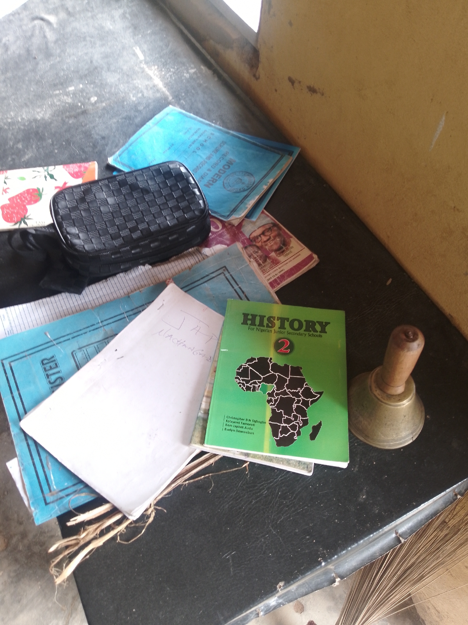 In Serti, Nigeria, a dark, textured surface, possibly a table, is covered with various items. A green book titled "HISTORY For Nigerian Junior Secondary Schools" with "2" prominently displayed is on the right, featuring a black outline map of Africa. Next to it, a brass handbell with a wooden handle rests on the surface. Several blue-covered books and papers are scattered around. One blue book is partially visible, displaying text and a circular seal. A white sheet of paper with handwritten notes is also present. To the left, a black woven pouch or bag lies on a patterned item, which appears to be a notebook with a strawberry design. A black cloth is draped over part of the bag. The background consists of a textured, light-colored wall and a dark, possibly tiled floor.