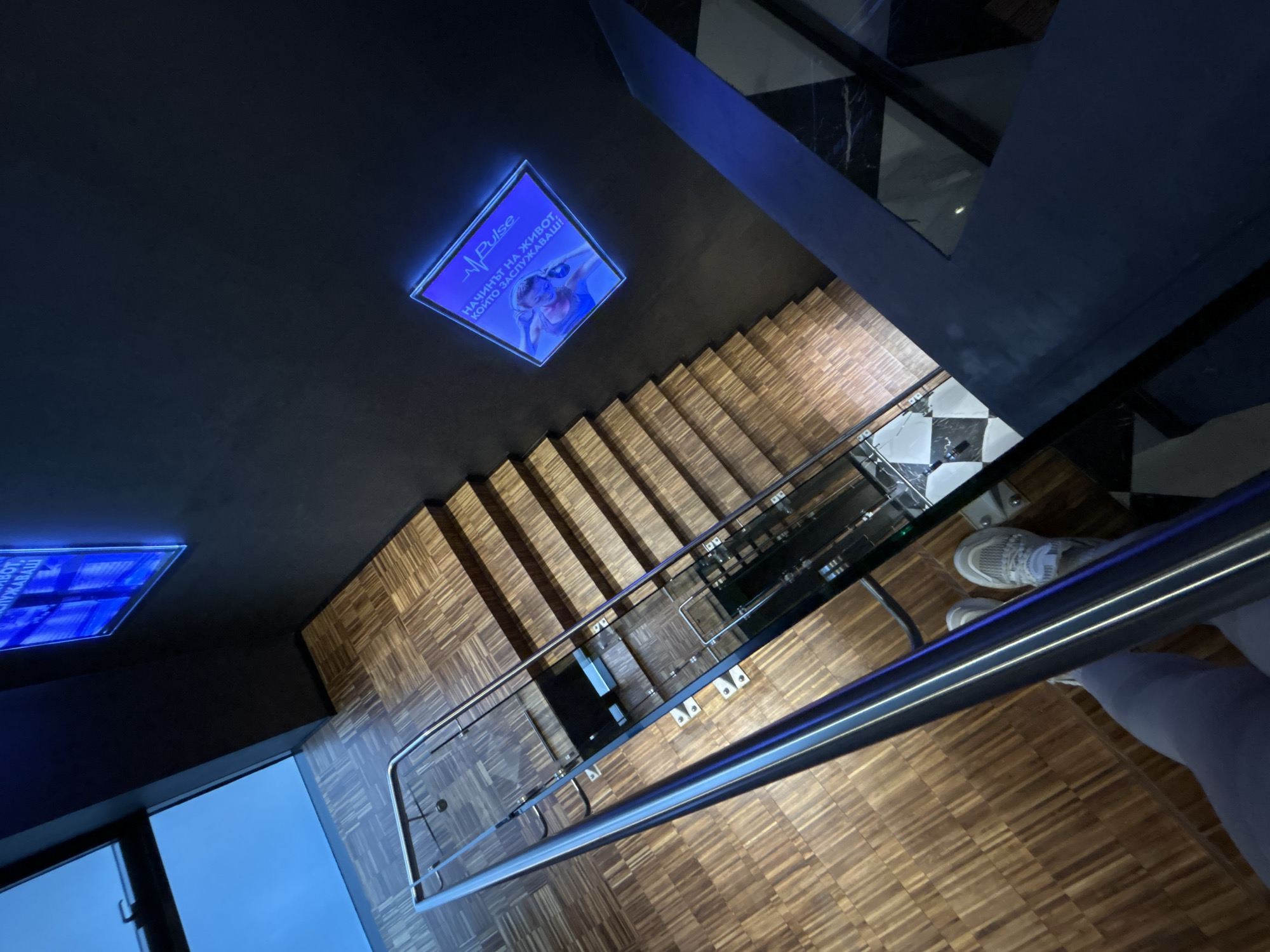 An overhead shot shows a staircase with wooden steps leading downwards. A metal railing runs along the right side of the staircase, and a person is visible in the bottom right corner, wearing white shoes and light gray pants. The ceiling above is dark, with two backlit signs. The top right corner shows a black and white tile pattern. The location is in the city of Plovdiv, Bulgaria.