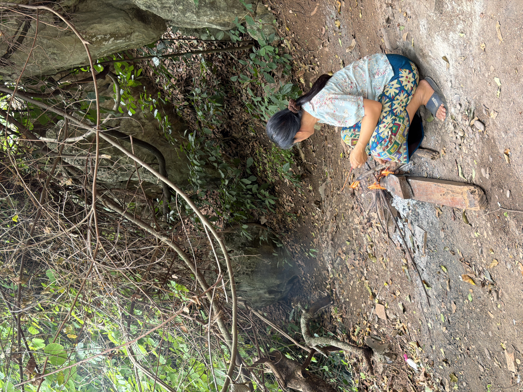 A woman with dark hair tied back with a brown scrunchie is crouched in a dirt clearing in a forested area, likely in Muangkeo, Laos. She is wearing a light blue, floral-patterned short-sleeved shirt and a colorful, patterned skirt. Her attention is focused on a small campfire in front of her, where she appears to be tending to the flames with a stick. The ground is covered in dirt, dry leaves, and twigs. Large rocks and dense vegetation, including vines and trees, surround the clearing. There is evidence of a small fire with smoldering embers and ash.