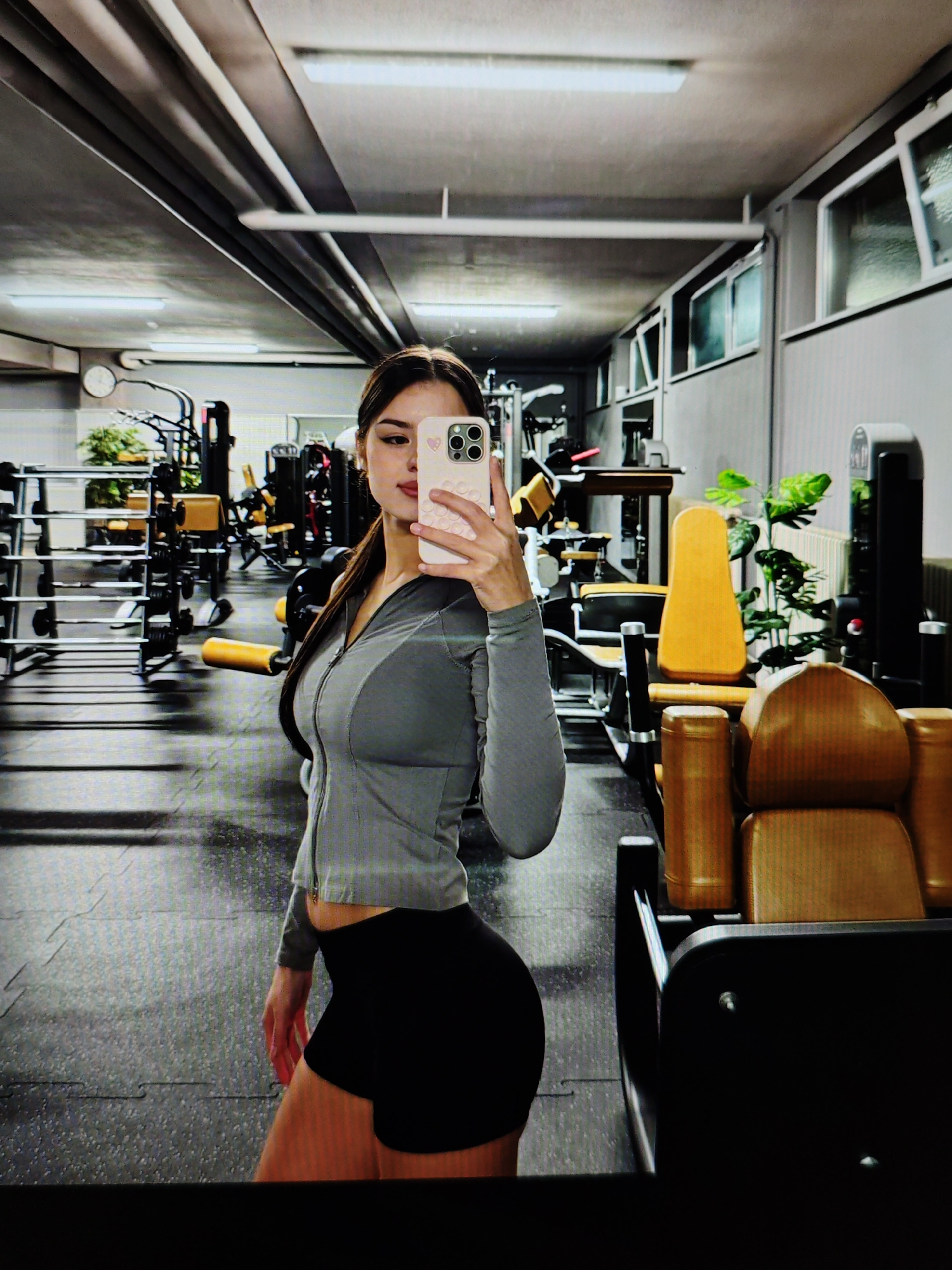 The image shows a young woman taking a selfie in a gym. She is wearing a grey zip-up jacket and black shorts. The gym appears to be well-equipped with various machines and weights. The lighting is bright, and the overall atmosphere suggests a modern fitness facility. The woman's reflection is visible in a mirror, capturing her pose and the surrounding gym environment. The image does not provide any specific clues or context to suggest it is located in Baku City, Azerbaijan.