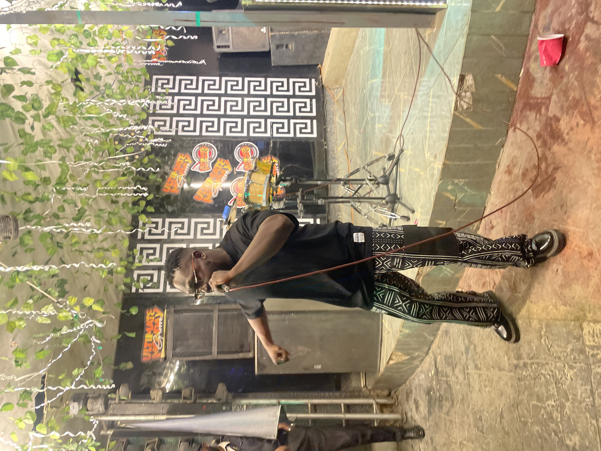 In Guzape, Nigeria, a Black male performer, wearing a black t-shirt and patterned pants, stands on a stage holding a microphone. He appears to be singing or speaking into the microphone, with his right arm extended and his fist clenched. The stage is adorned with artificial greenery and hanging white streamers, creating a festive atmosphere. Behind the performer, a drum set is visible, along with speakers and a black backdrop featuring a geometric pattern. To the left of the stage, another individual, likely a security guard, stands dressed in a dark uniform. The floor of the venue is tiled with a mixture of green and brown hues, and a red cup is lying on its side on the ground near the edge of the stage. The scene suggests an indoor event or performance space.