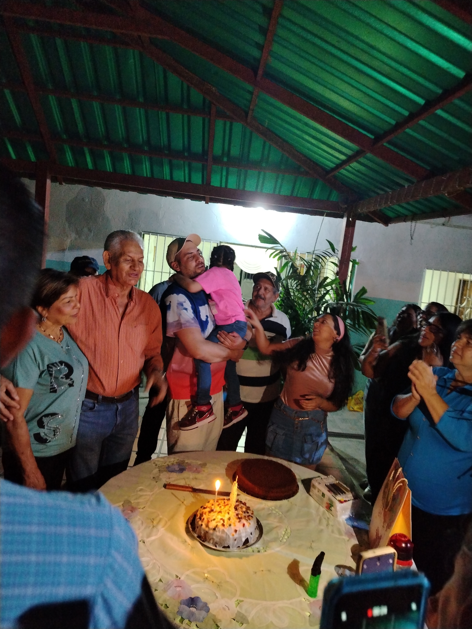 A group of people are gathered around a table with a birthday cake in Tinaquillo, Venezuela. The cake has a lit candle on it. A man is holding a child, and a woman is reaching towards them. Other people are standing around, some clapping their hands. There are other cakes, presents, and items on the table. The scene takes place in an open covered area with a green roof. The walls are white, and there is a plant in the background. A person is holding a smartphone to take a picture.