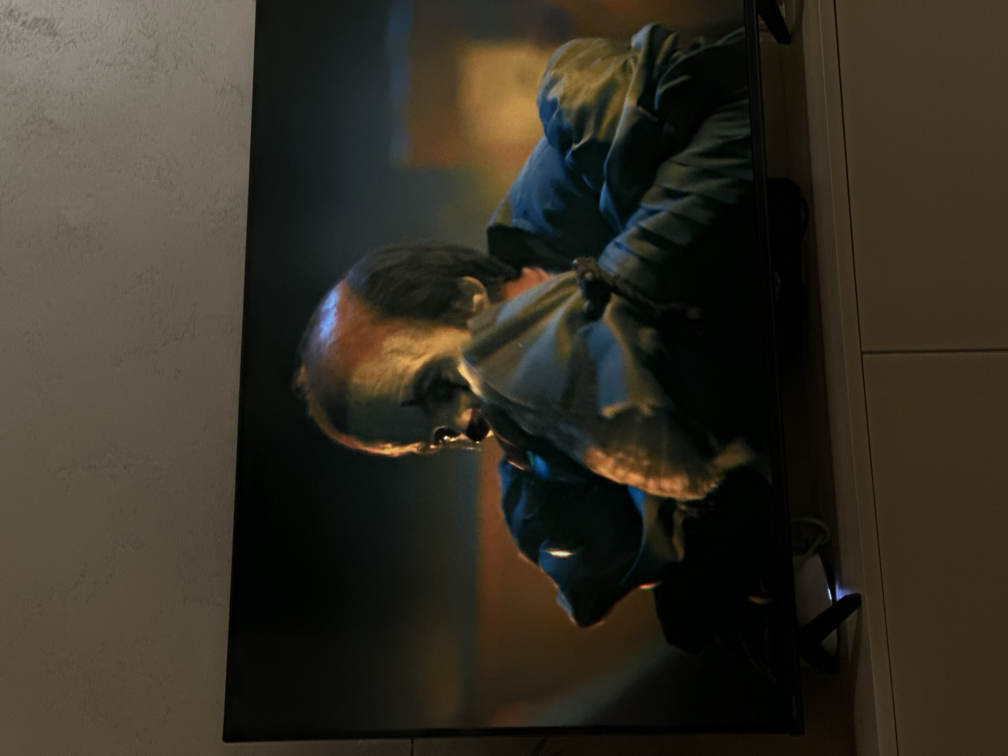 The image shows a television screen displaying a man in a dark, intense scene. The man has a partially bald head and is wearing a dark jacket or coat. He appears to be struggling, possibly bound or held. The background of the scene on the screen is dark and blurry, with hints of yellow and brown tones. The television is mounted on a wall next to a light-colored surface. The image provides no information to associate it with Sofia, Bulgaria.