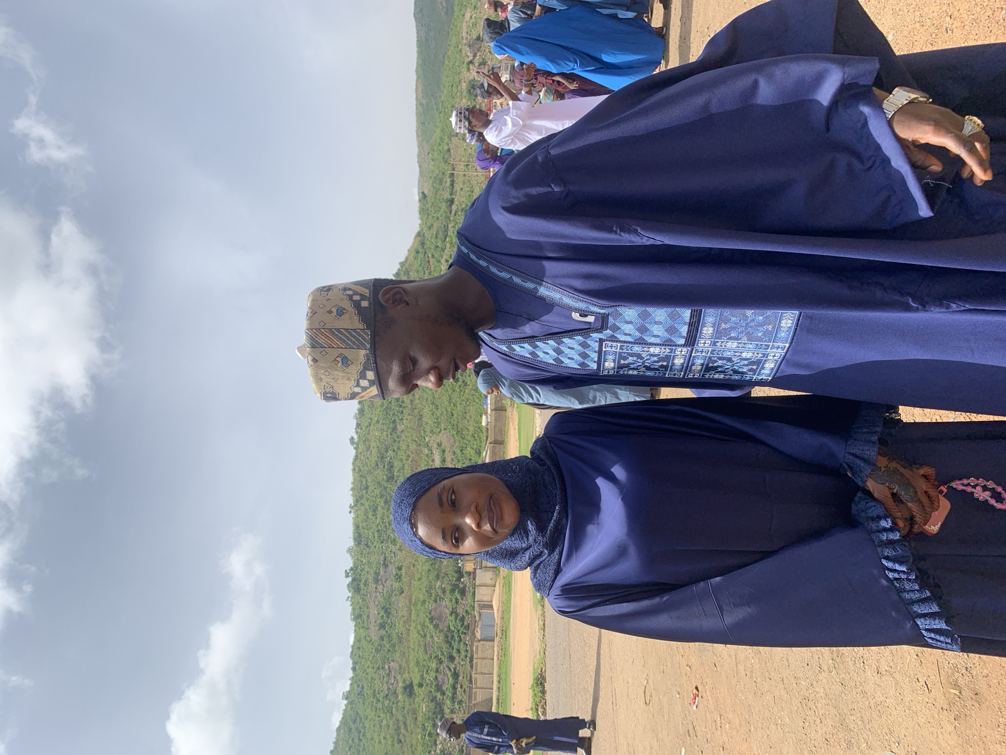 The image shows a man and a woman posing outdoors. The man wears a dark blue robe with patterned embroidery and a patterned kufi hat. He has a watch on his left wrist. The woman is wearing a dark blue hijab and robe. Behind them is a green hillside and a cloudy sky. Other people are visible in the background. They are located in the city of Naraguta Hausawa, Nigeria, as indicated by the location data.