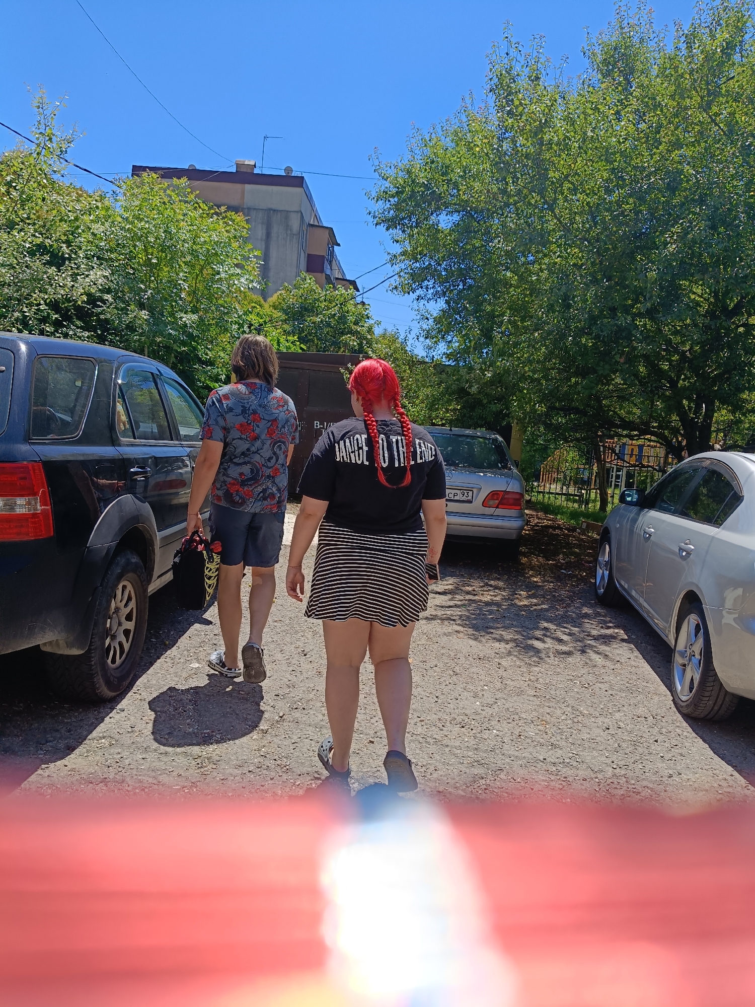 In this outdoor shot, two individuals walk away from the camera on a sunny day. The person on the left, with medium-length brown hair, wears a blue t-shirt with a red and black floral pattern and grey shorts. They are carrying a black bag with yellow string. The person on the right has bright red hair styled in two braids and wears a black t-shirt with white text that reads "DANCE TO THE END" and a black and white striped skirt. They are wearing black and white patterned slides.
They are walking on a gravel path between two parked cars. To the left is a black SUV, and to the right are two silver sedans. In the background, there are lush green trees, a building with a beige facade, and a blue sky. A wire hangs across the sky, and a small antenna is visible on the building.
The scene appears to be a residential or semi-rural area. The bright sunlight suggests it is midday. The overall mood is casual and ordinary, with no strong emotions conveyed. The visible text on the t-shirt is the only discernible text in the image. The location is identified as Oryol-Izumrud, Russia.